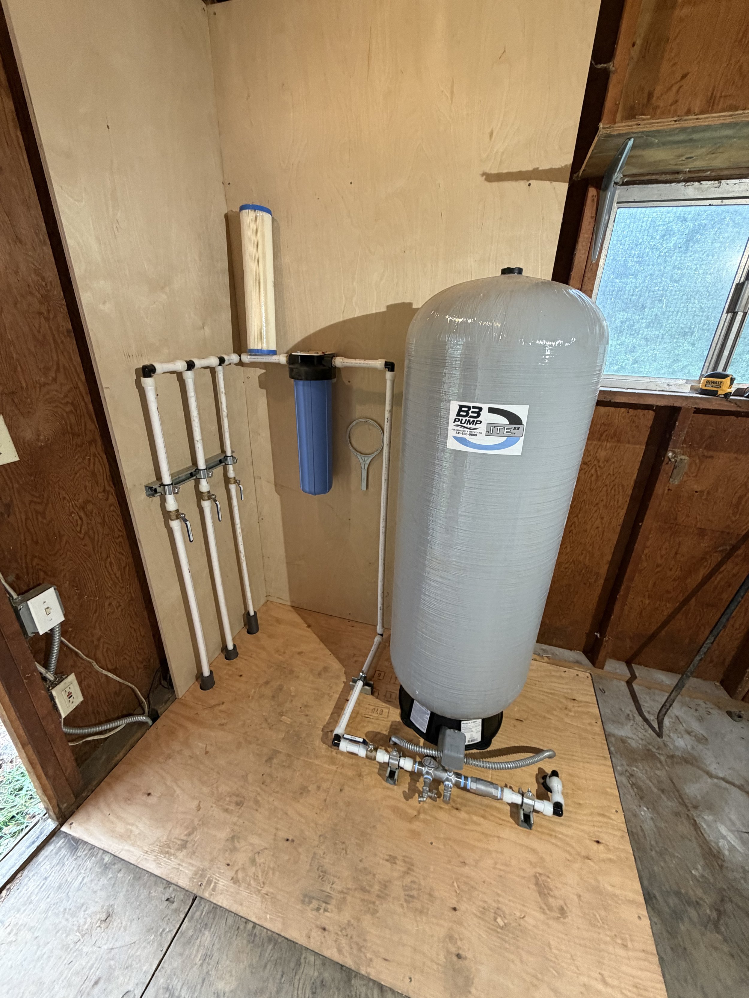 Installation of water filtration system with pipes, large tank, and filter components in a wooden room with a window.