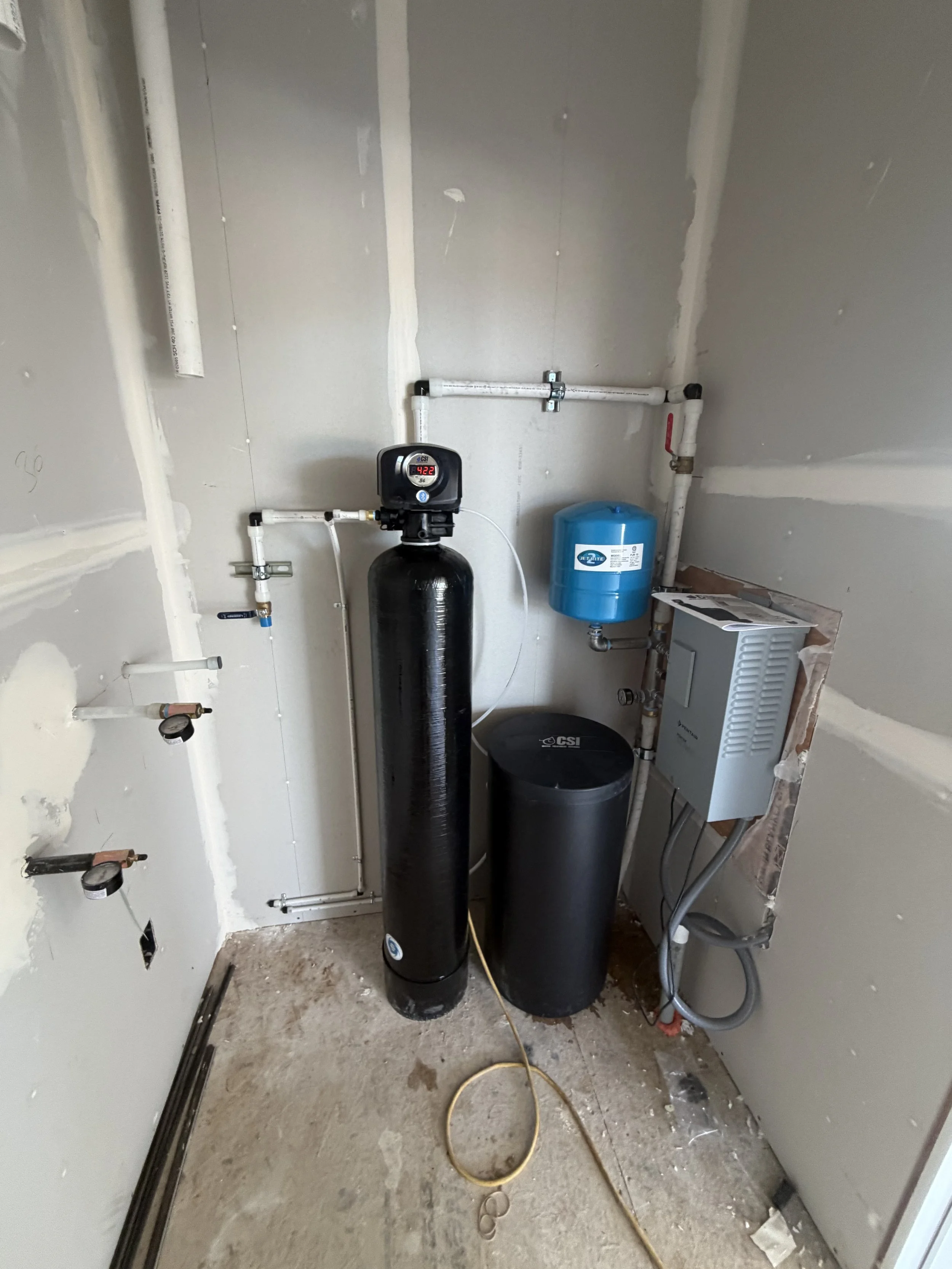 A utility room under construction with water filtration and pump equipment, including pipes, and a blue pressure tank.