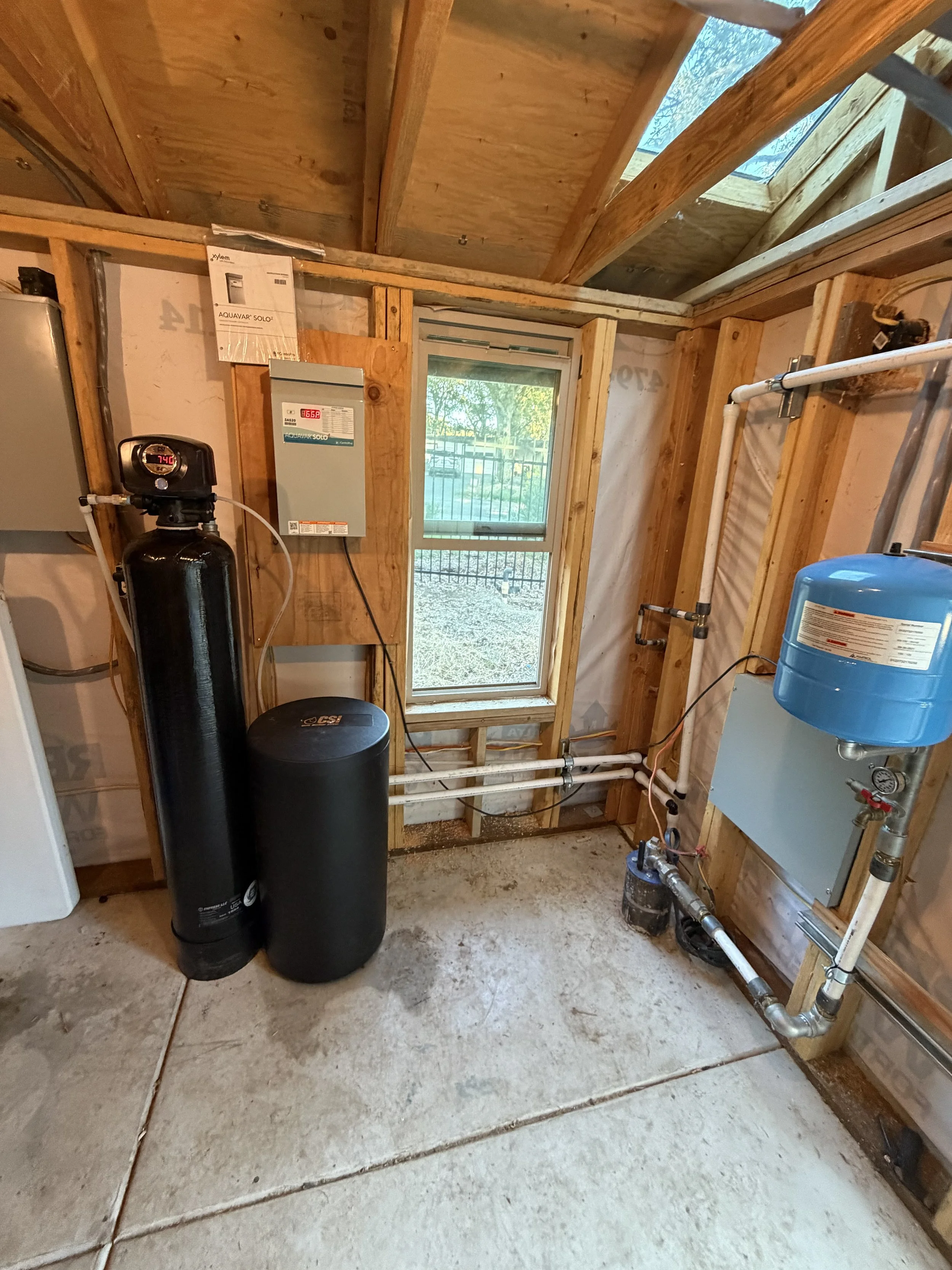 A utility room with wooden framing, a window, and several water pipes. There is a black water softener tank and a blue water filtration system installed on the right side. An electrical panel and a water heater are also visible.