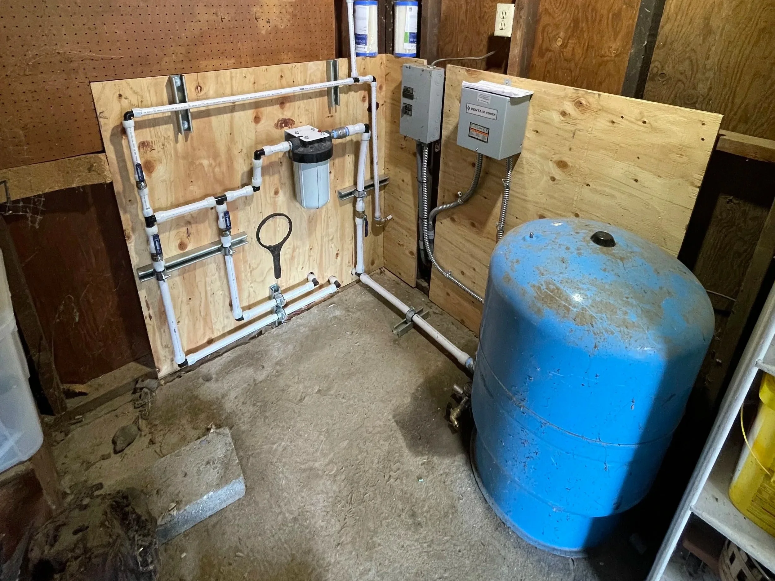 A water well pressure tank and filtration system in a basement or utility room with exposed plumbing and electrical connections.
