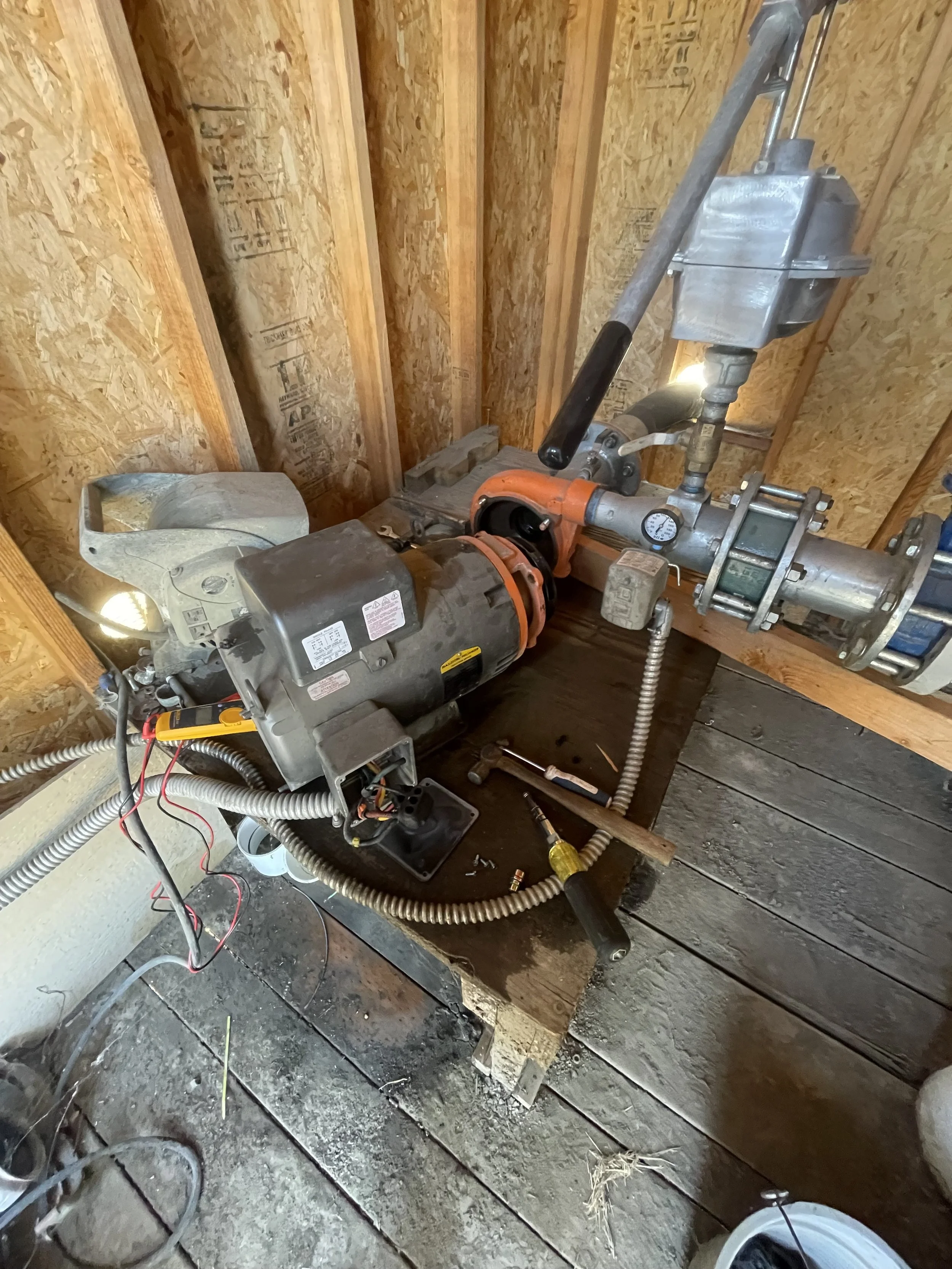 A irrigation water pump system installed inside a wooden structure with an electric motor, pipes, gauges, and tools nearby.