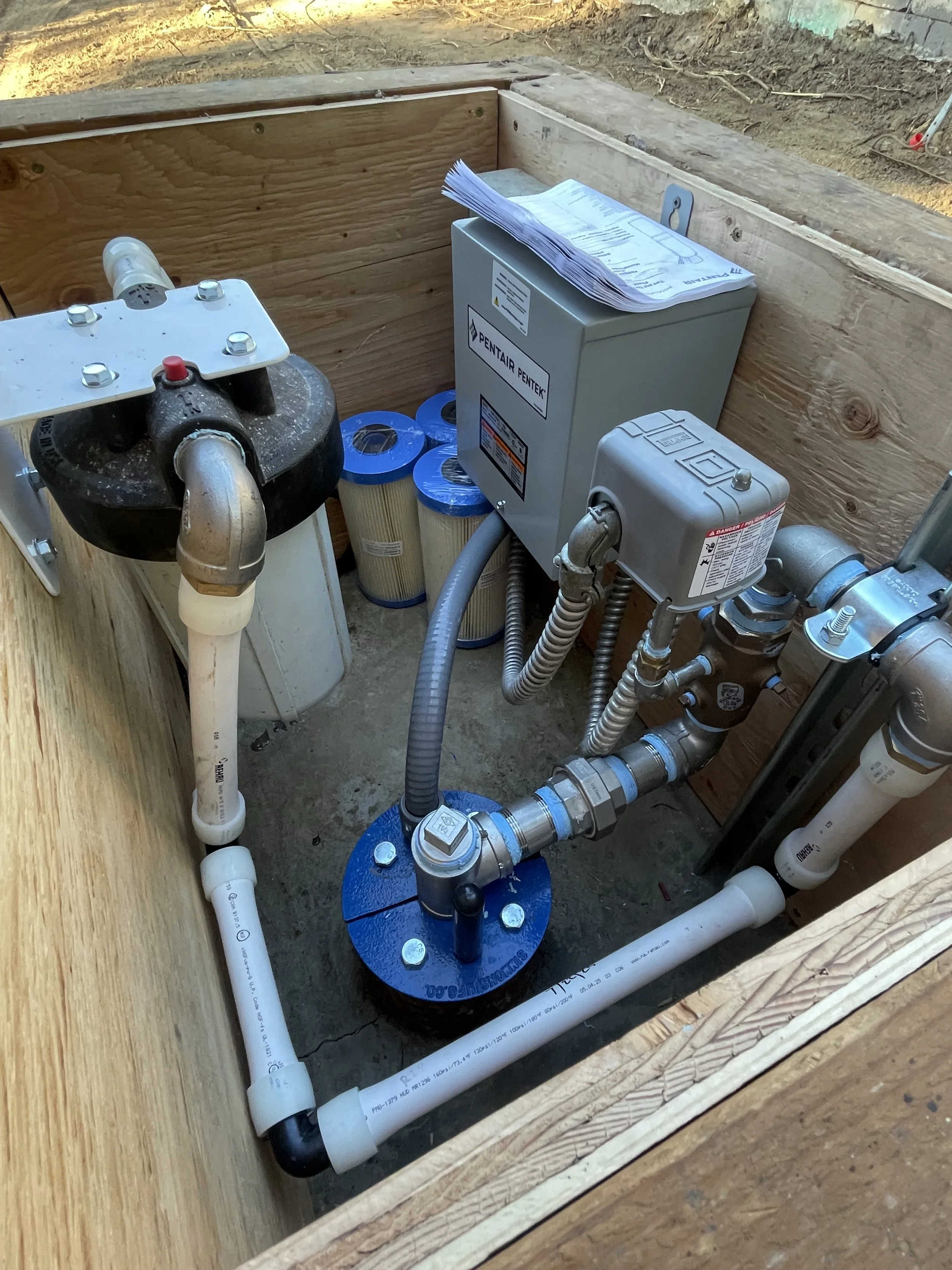 This image shows a small well pump system with filters, piping, and electrical components installed inside a wooden enclosure at a construction site.