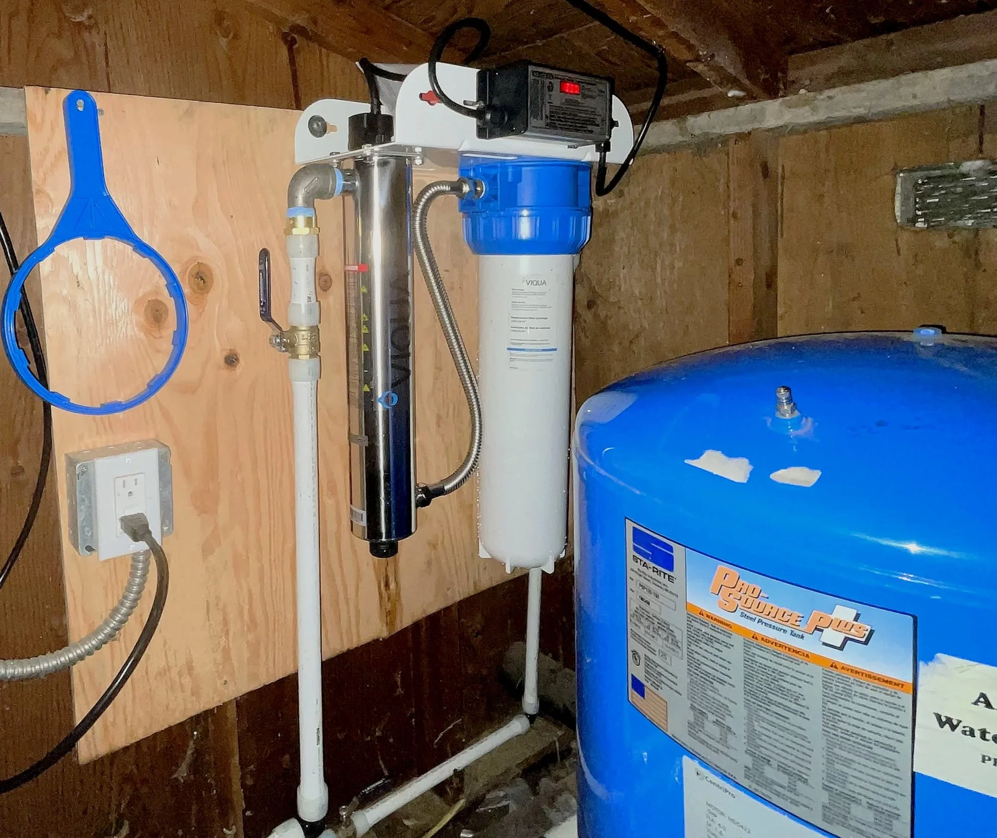 A uv water filtration system and air pressure tank installed on a wooden wall in a utility room.