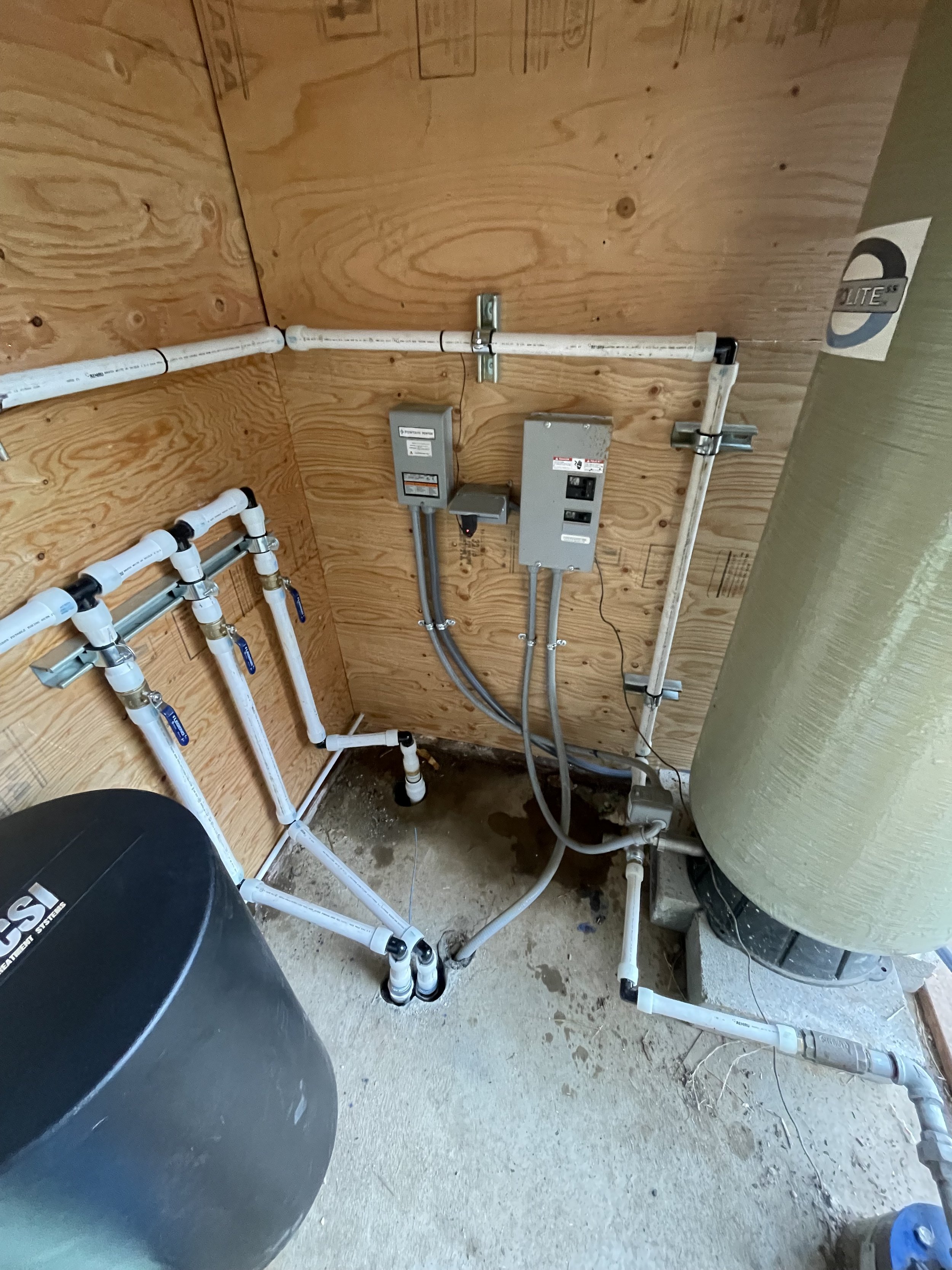 water well control box, Piping and electrical setup in a pump room with wood walls, a water pressure tank, and electrical control boxes.