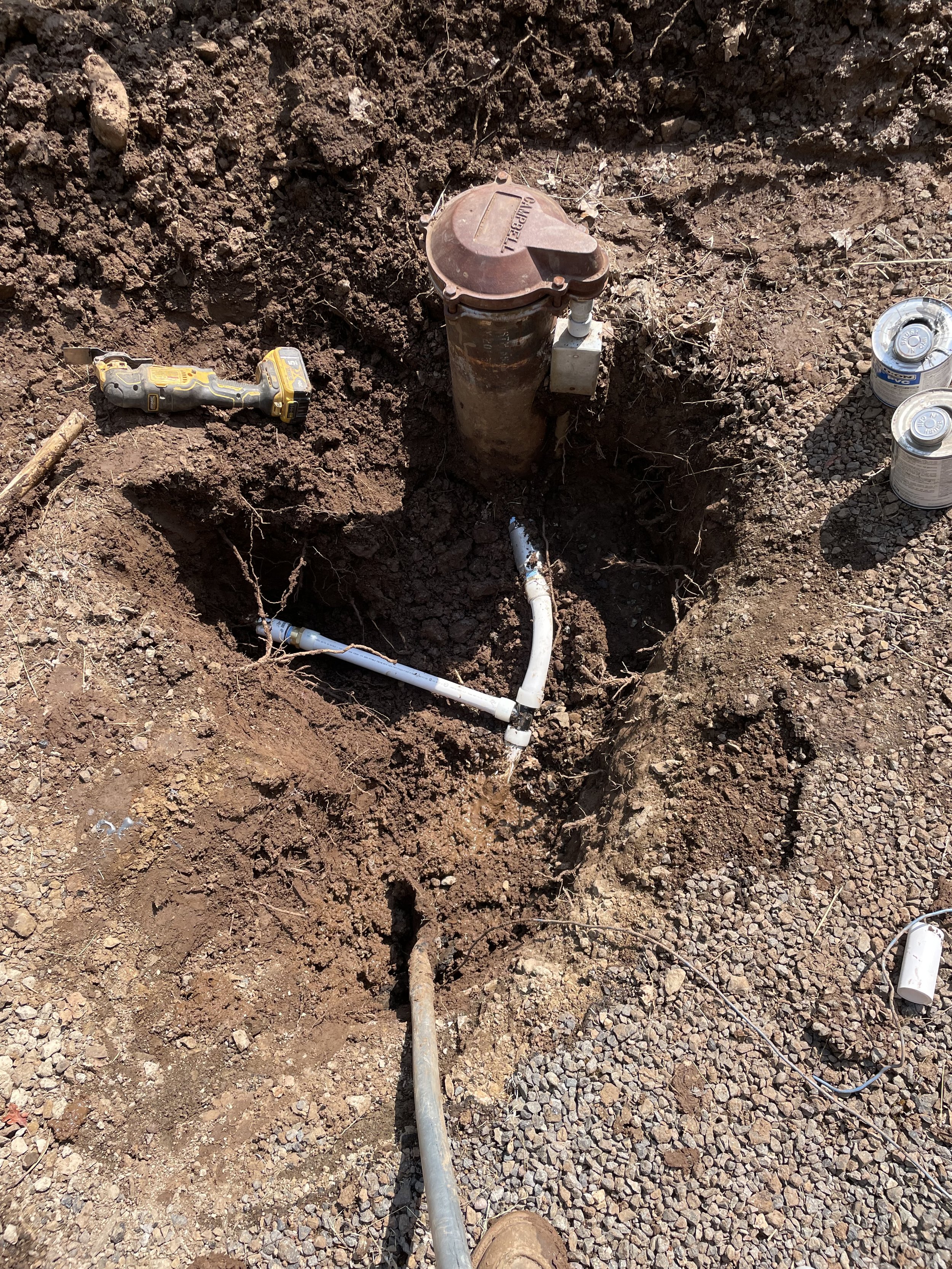 Digging site with plumbing installation, including PVC pipes, exposed in the ground, with tools and canisters nearby.