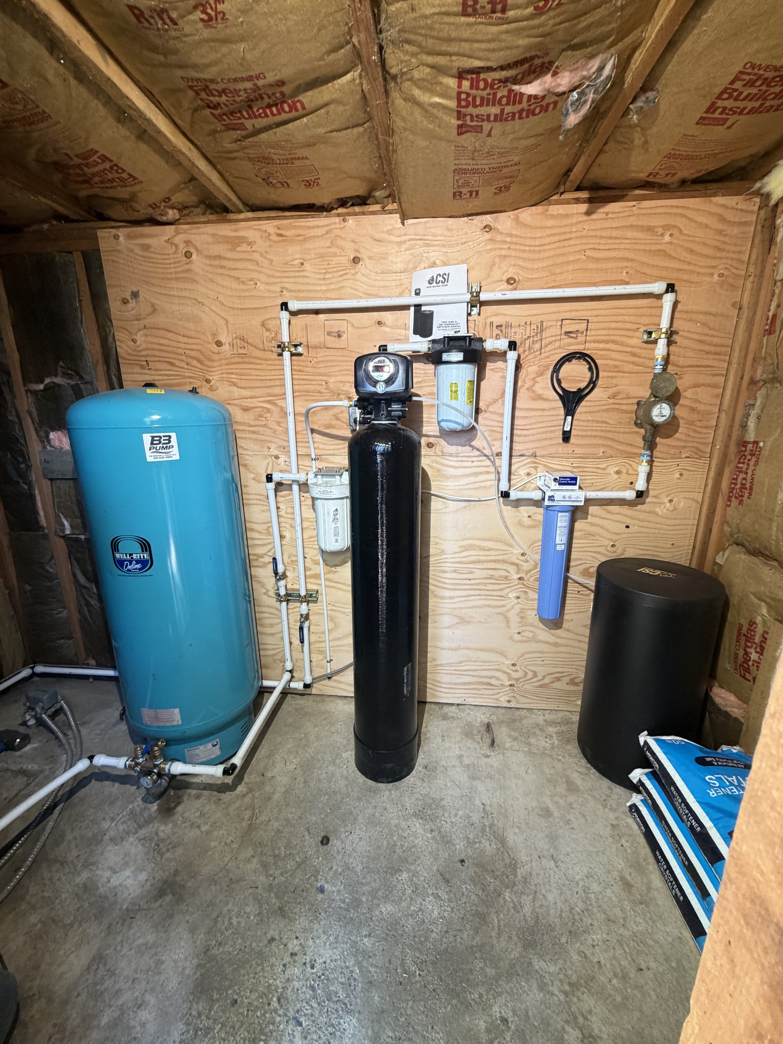 A water softener system with a black tank, a blue tank, and various pipes and gauges in a utility room or basement.