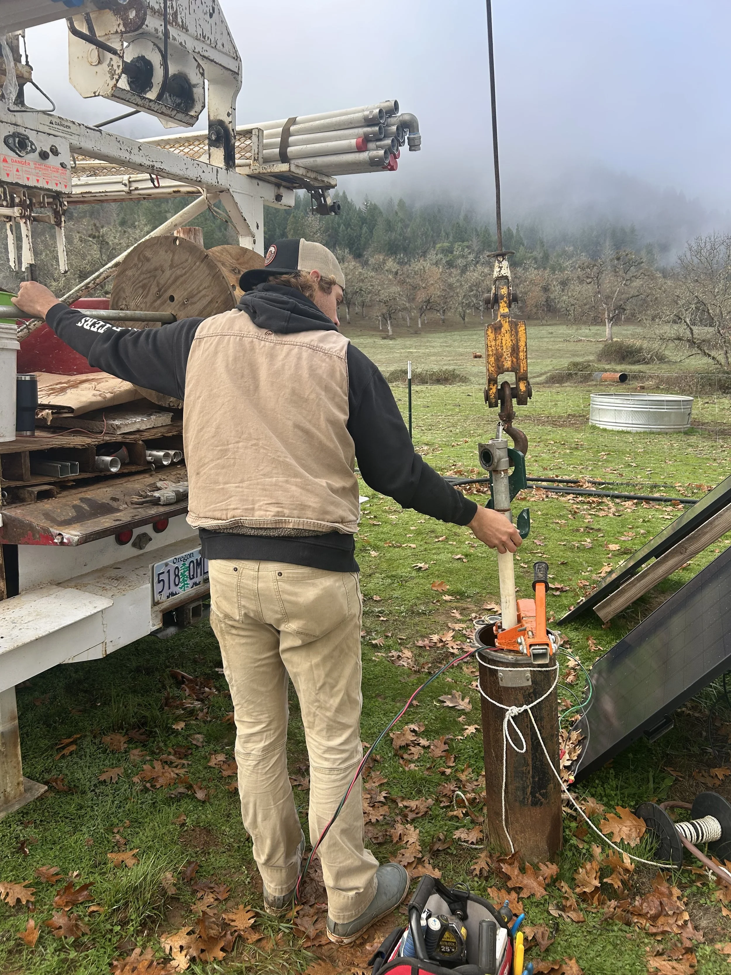 A person is pulling a well pump with a pump hoist, wearing a beige vest and beige pants, on a grassy area with fallen leaves, with equipment, a truck, and a misty background of trees and hills.