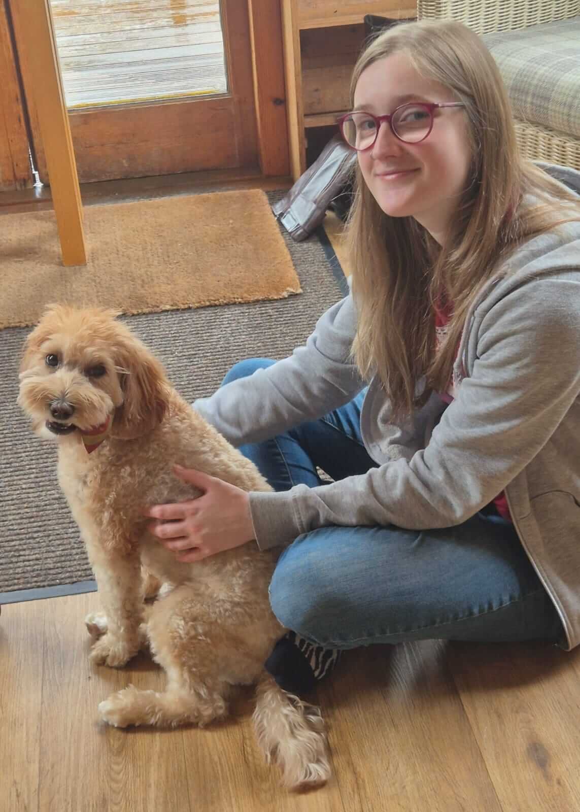 A young woman is dog sitting for a golden curly-haired small dog indoors near a door with a window.