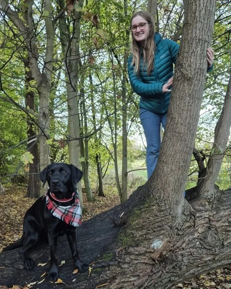 A dog walker in Edinburgh, peeking out from behind a tree trunk in a wooded area, with a black Labrador dog sitting on the fallen tree near her; the dog is wearing a red and white plaid bandana.