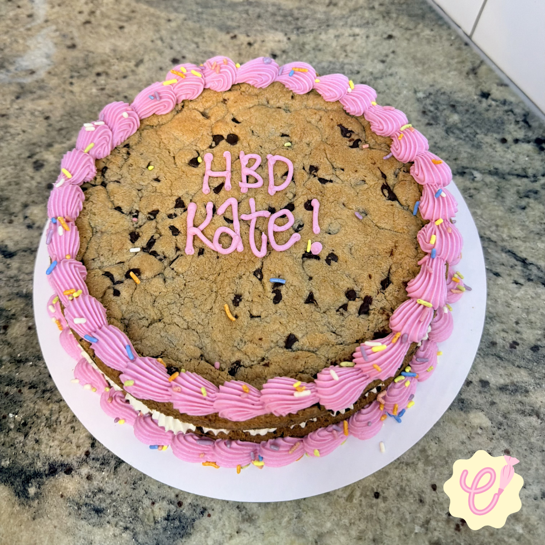 A large chocolate chip cookie cake decorated with pink icing and rainbow sprinkles. Pink icing writes 'HBD Kate!' in the center.