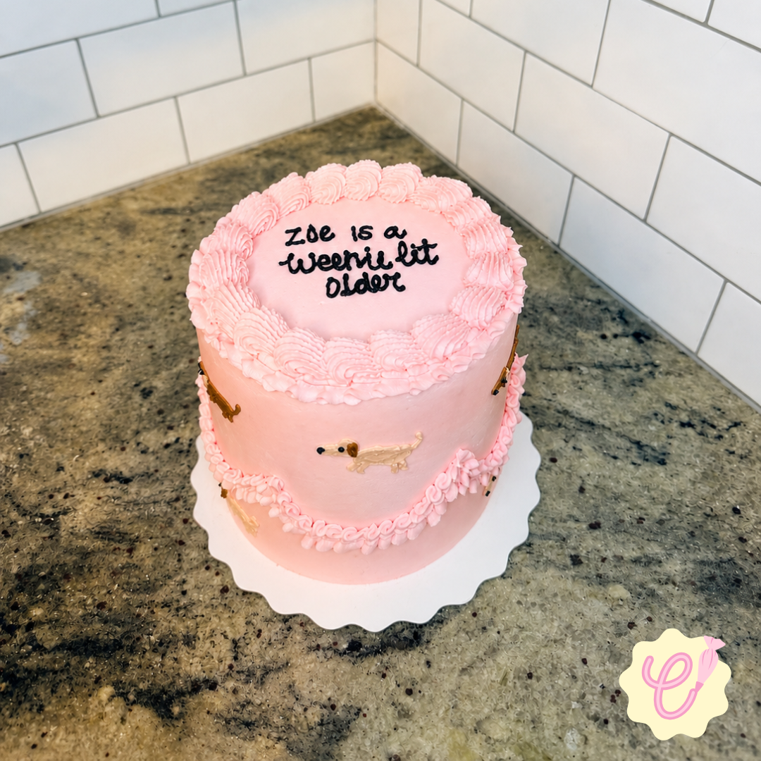 A pink birthday cake with a border of piped frosting, decorated with small dachshund dog figurines. The top has a message written in black icing: 'Zoe is a weenie bit older.'