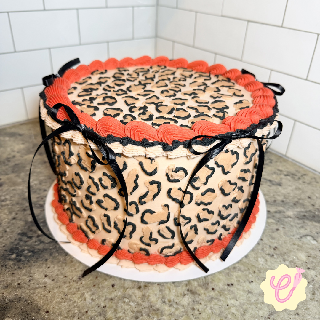 Leopard print cake with pink and black piping, black ribbon bows, placed on a white cake board on a granite countertop with white tiled wall in the background.