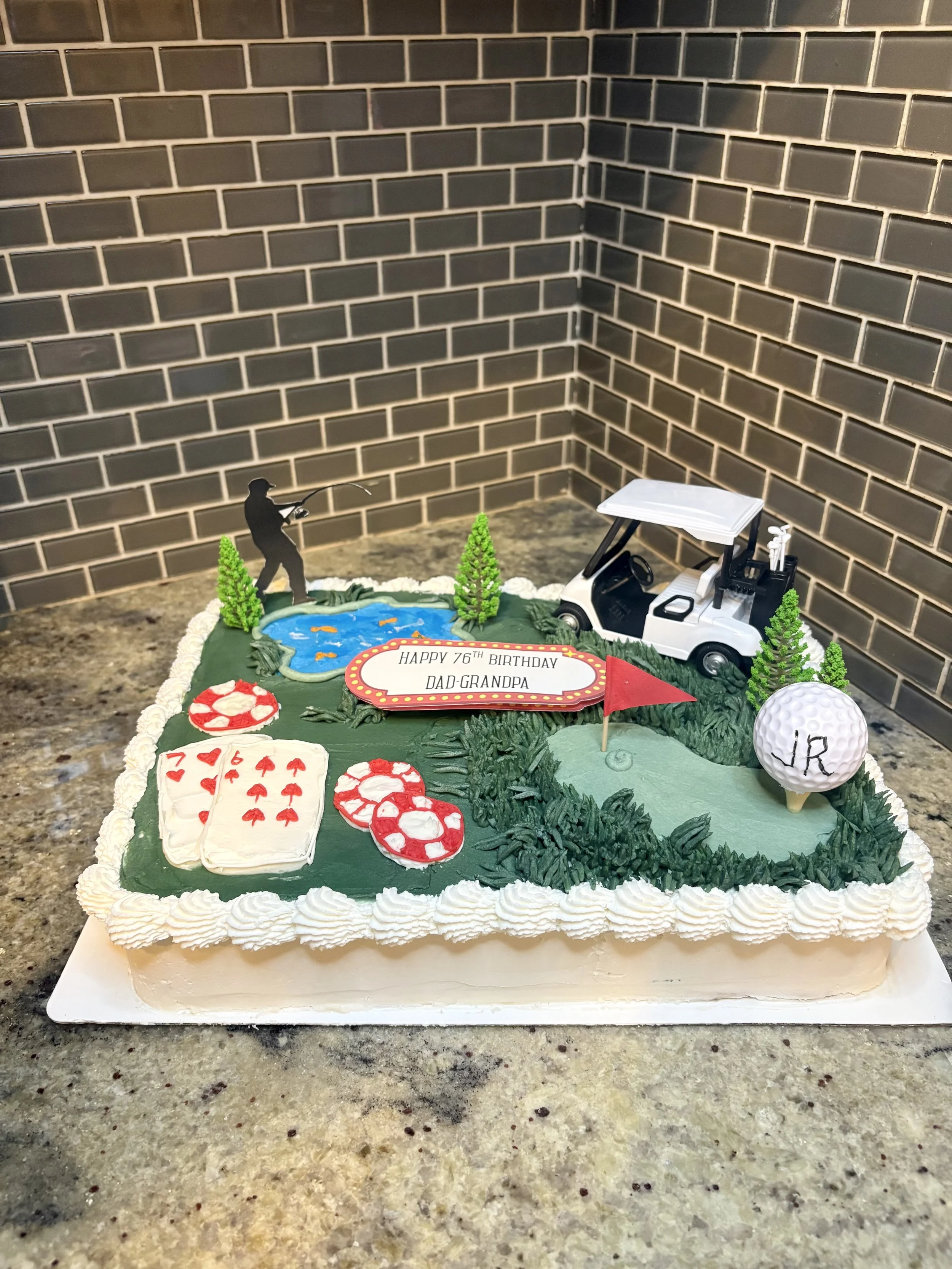 A golf-themed birthday cake decorated with a miniature golf course, a golf cart, a golfer figure, a small pond, trees, a red flag with the message 'Happy 76th Birthday Dad-Grandpa,' a golf ball with 'JR' written on it, and playing cards.