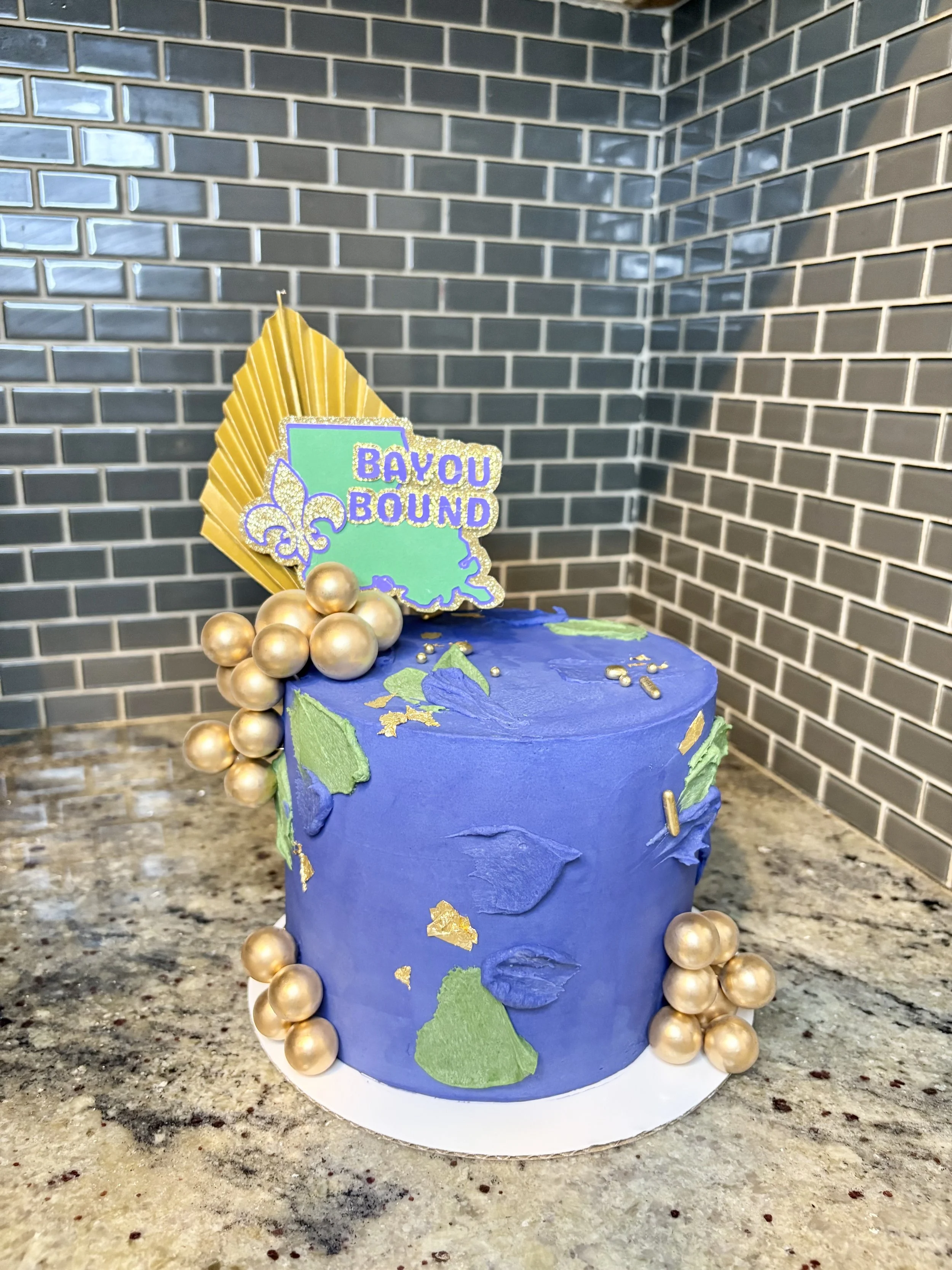 A decorated cake with blue and green themed decorations, gold accents, and a topper that says 'Bayou Bound' with a Louisiana map and fleur-de-lis symbol.