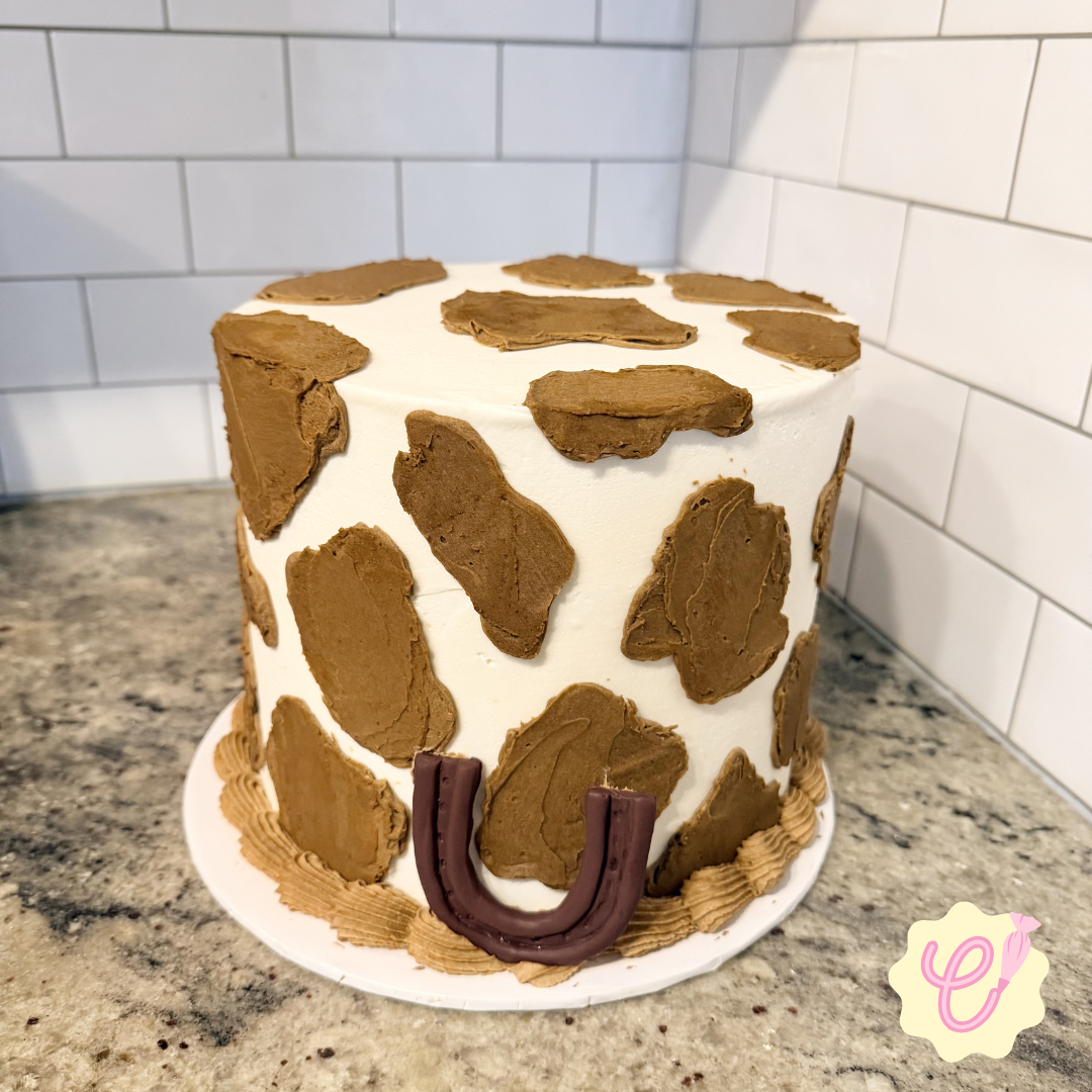 A cake decorated with chocolate frosting patches and a horseshoe shaped decoration on the front, sitting on a marble countertop against a white tiled wall.