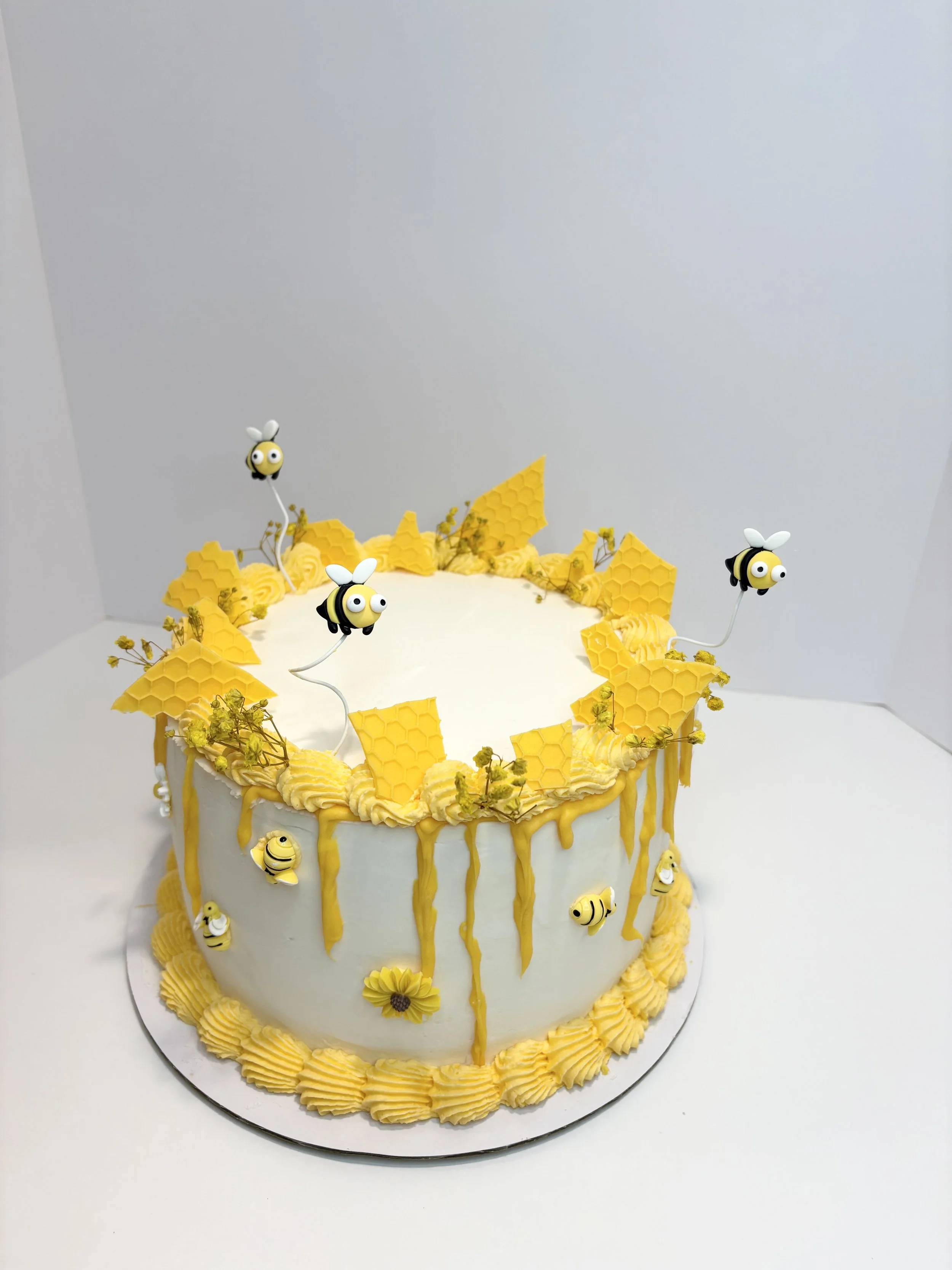 A white cake decorated with yellow honeycomb accents, bees, and honey dripping decor, placed on a silver cake board.