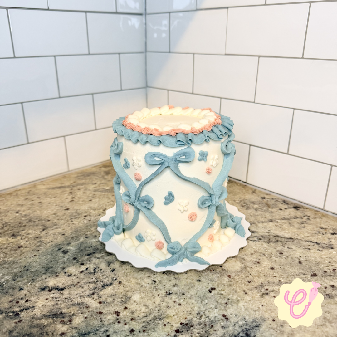 A tiered cake with white frosting decorated with blue and pink ribbon-like icing and small white, pink, and blue floral designs, placed on a cake board on a granite countertop.