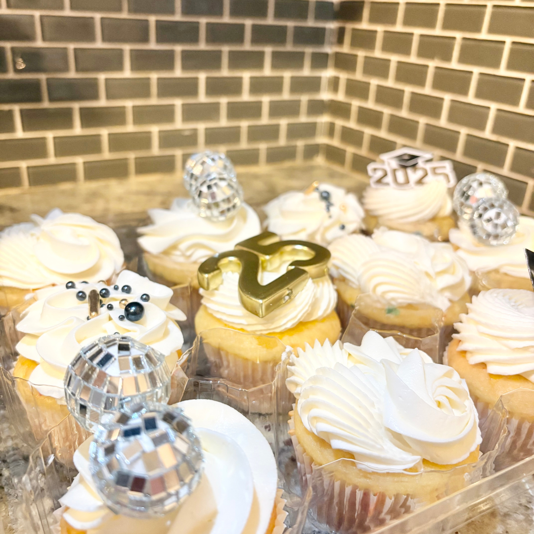 A tray of decorated cupcakes with white frosting, disco ball toppers, a gold number 2, and a graduation cap and year '2023' decorations, set against a brick wall background.