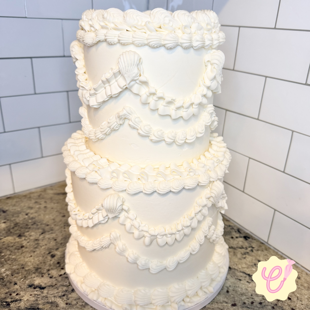 A four-tiered white cake decorated with shell-shaped and piped frosting accents, placed on a granite countertop against a white tiled wall.