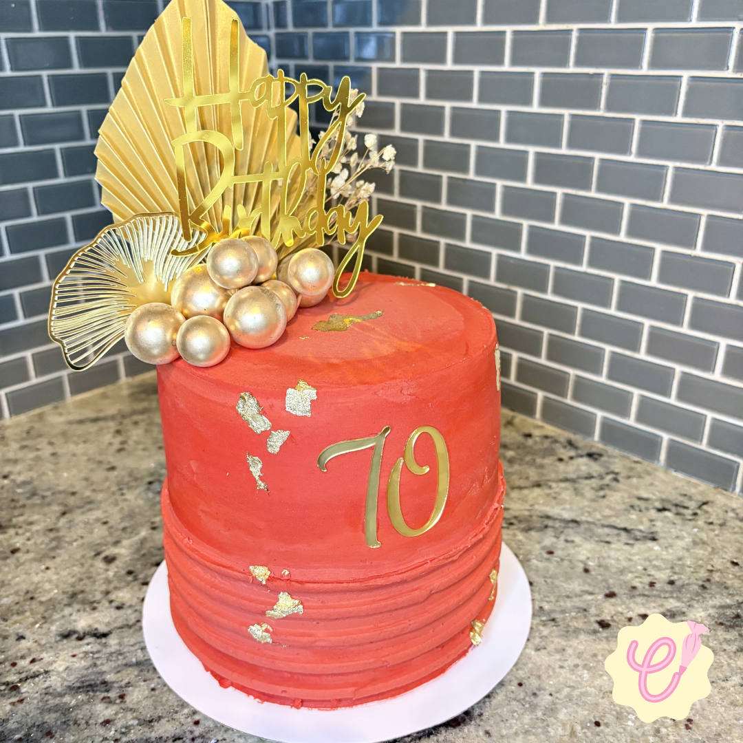 A red birthday cake decorated with gold accents, topped with a cluster of pearl-like balloons, a gold "Happy Birthday" topper, and a gold number 10 on the side, placed on a white cake board on a granite countertop with a gray brick wall in the backgr