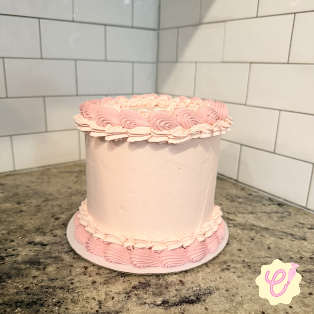 A round pink cake with decorative pink and white frosting on top and around the base sits on a granite countertop in a kitchen with white subway tile backsplash.