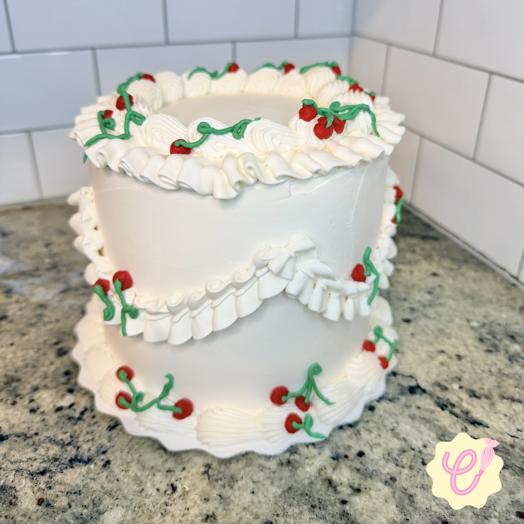 White two-tier birthday cake decorated with red cherries and green icing in a holiday pattern, placed on a gray speckled countertop against a tiled wall background.