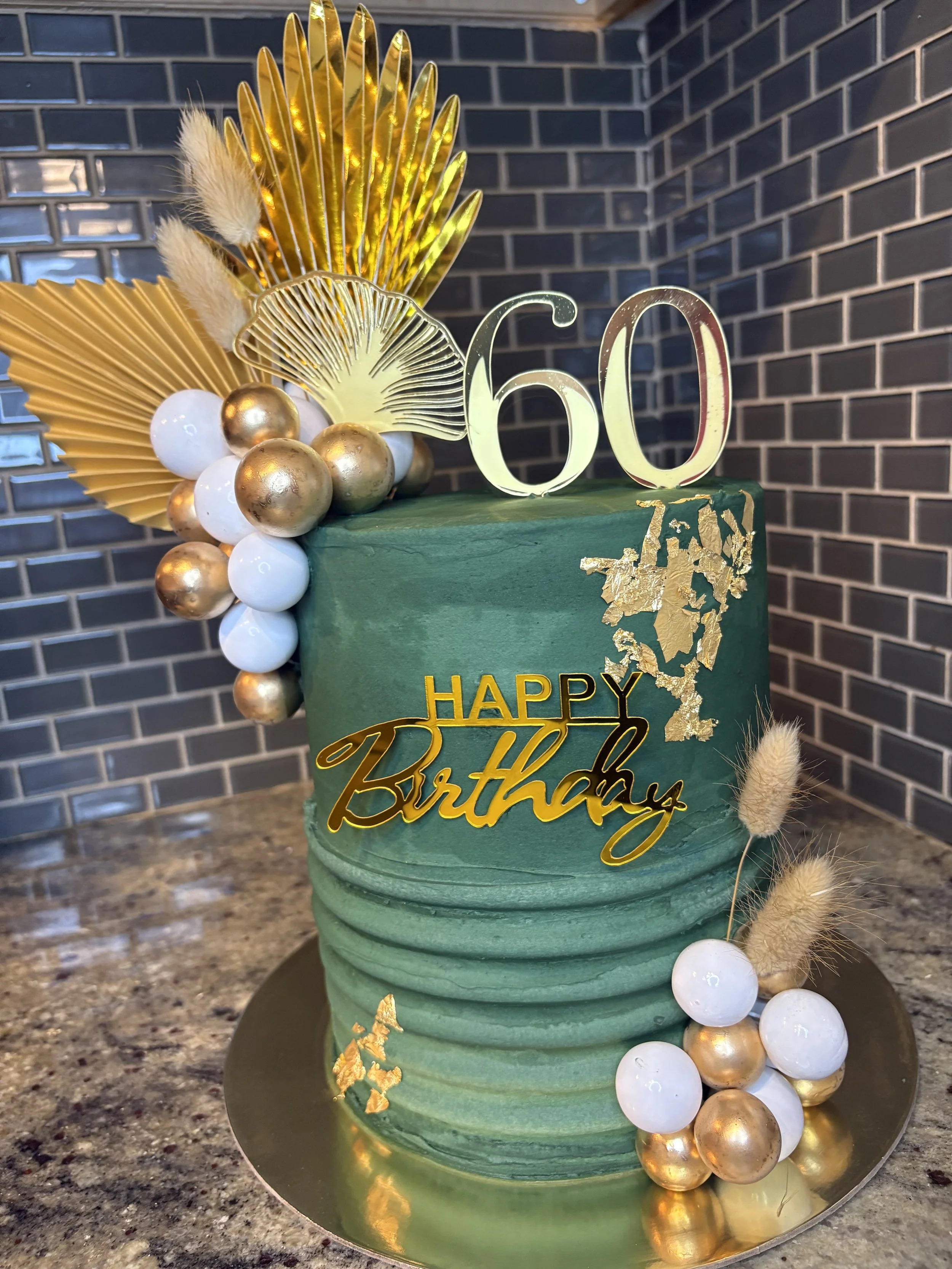 A green birthday cake decorated with gold and white ornaments, including a "60" topper, gold leaves, and "Happy Birthday" written in gold. The cake has textured ridges and is placed on a golden cake board.