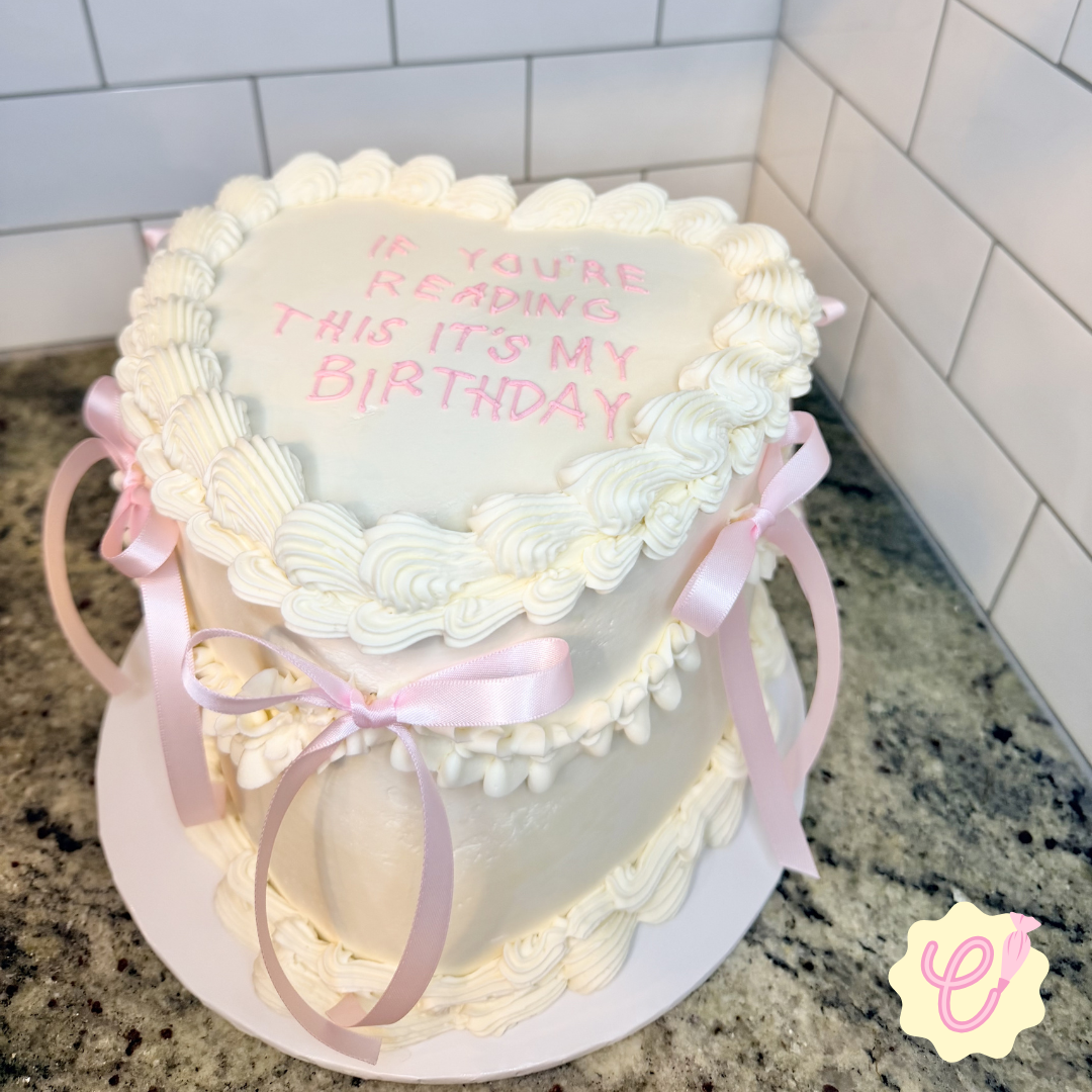 A heart-shaped birthday cake decorated with white icing, pink ribbons, and pink lettering that reads, "If you're reading this it’s my birthday."