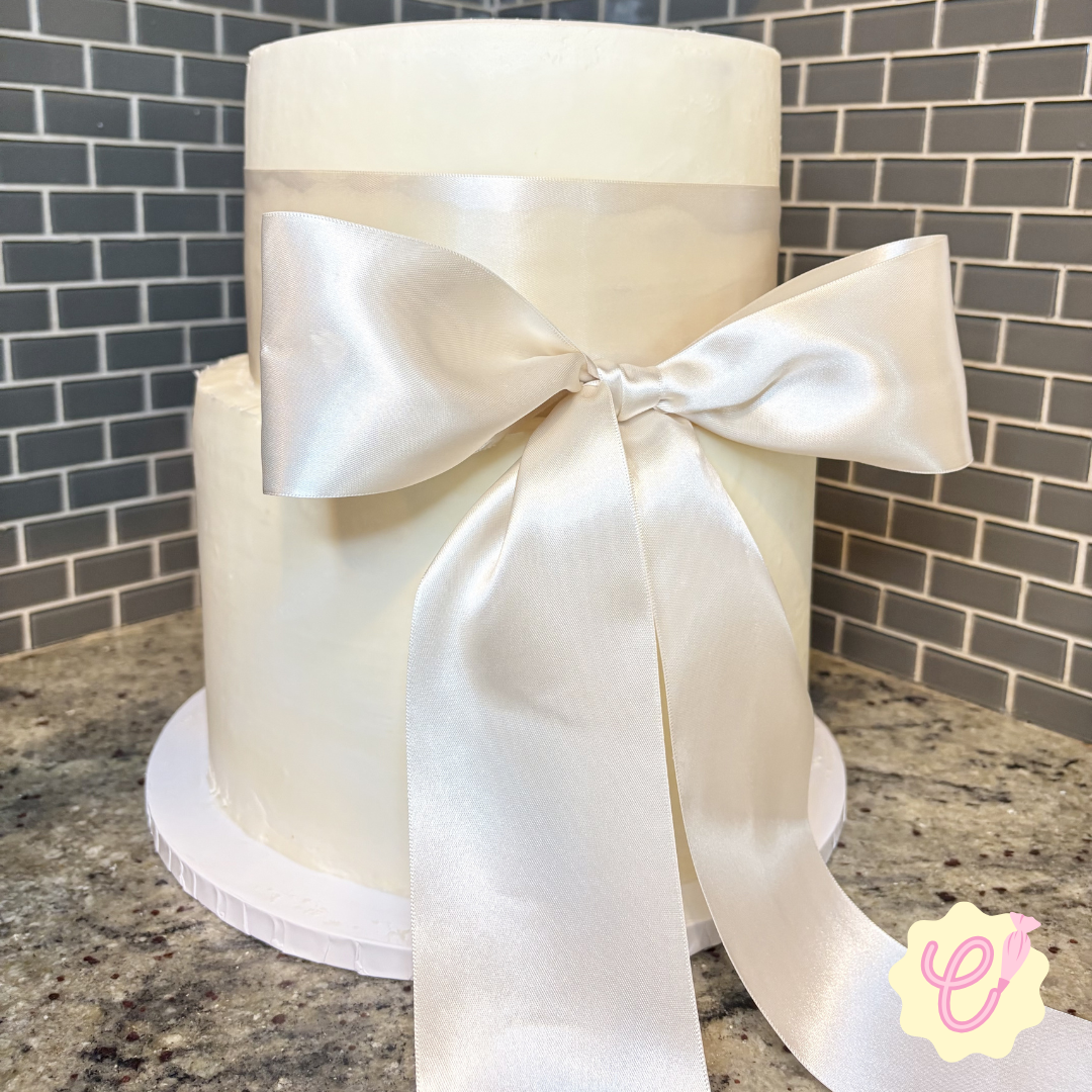 Two white wedding cakes stacked with a satin ribbon and bow on the front, placed on a granite countertop with a gray brick wall background.