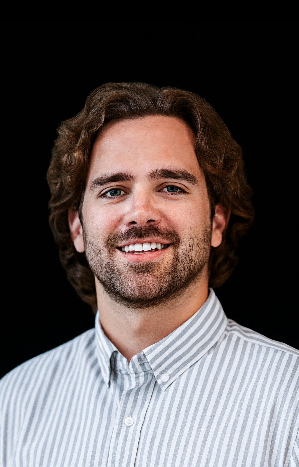 A young man with wavy brown hair, blue eyes, and a beard, wearing a light blue and white striped button-up shirt, smiling against a black background.