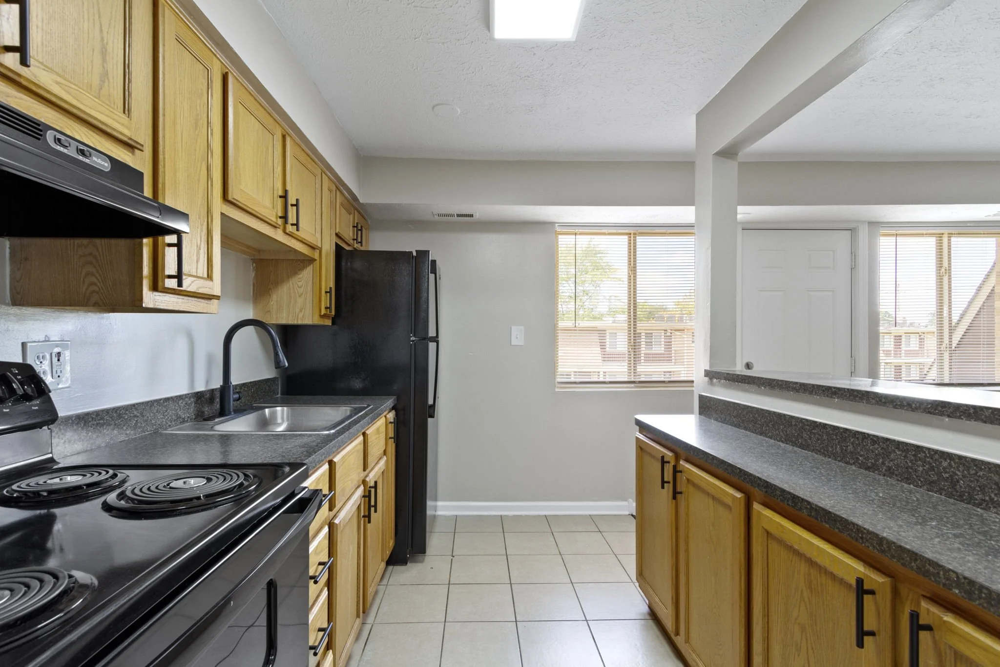 A kitchen with wooden cabinets, black appliances, a gray countertops, a double sink, and windows with blinds overlooking neighboring buildings.