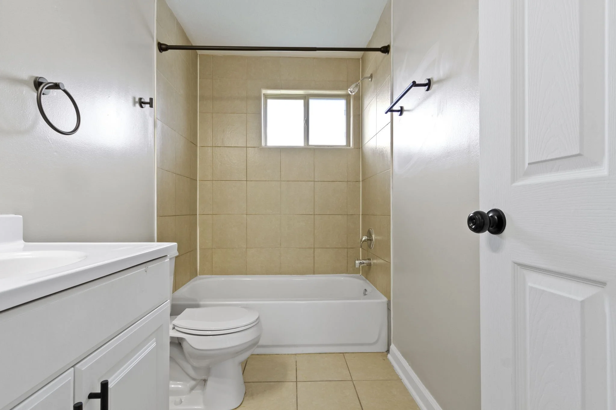 Bathroom with beige tiled walls, small window, white bathtub, toilet, and white vanity, with black fixtures and a door slightly open.