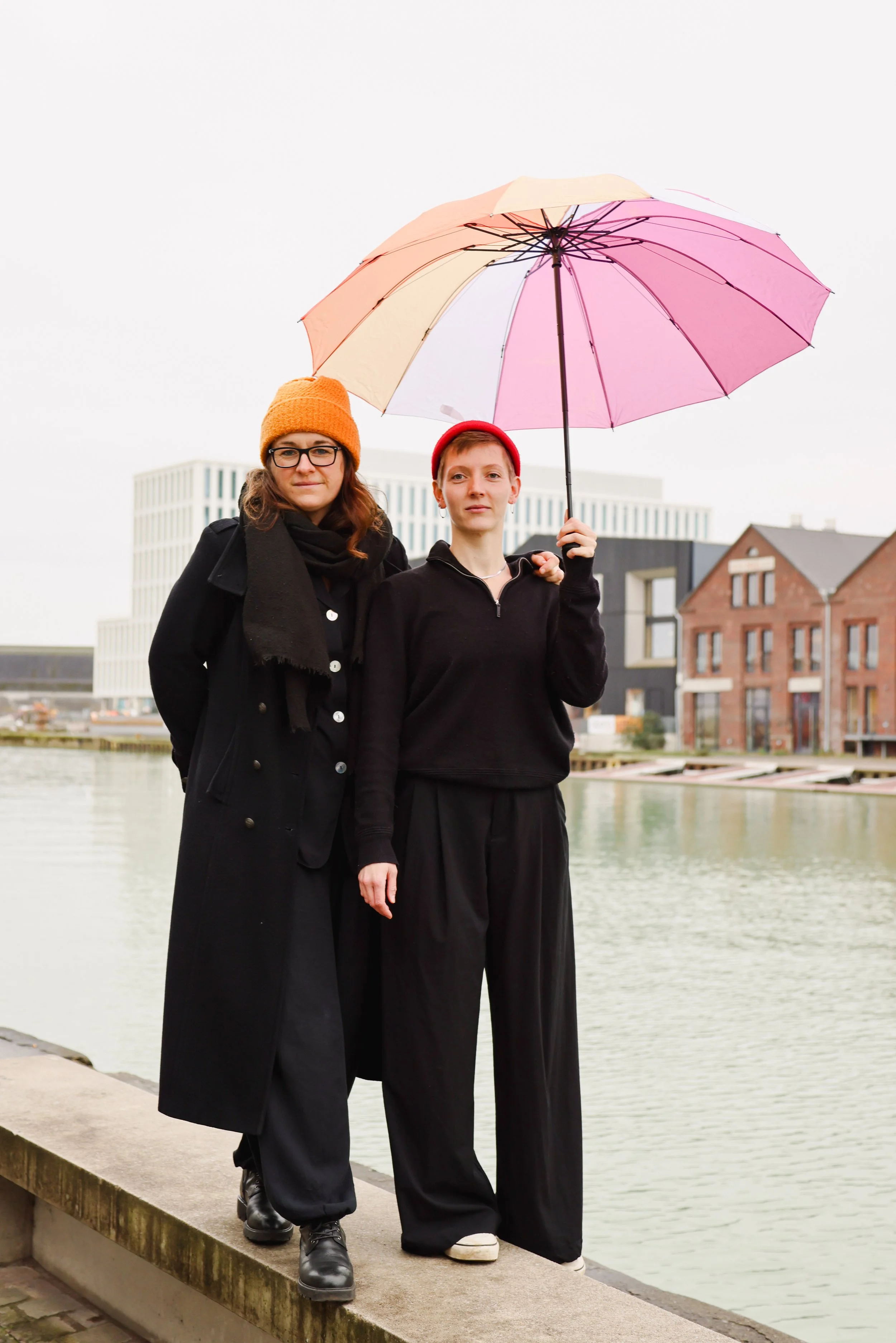 HANNA&LIA in schwarz gekleidet mit bunten Mützen untem einem lesbian pride Regenschirm, stehend, im Hintergrund Wasser und Häuser