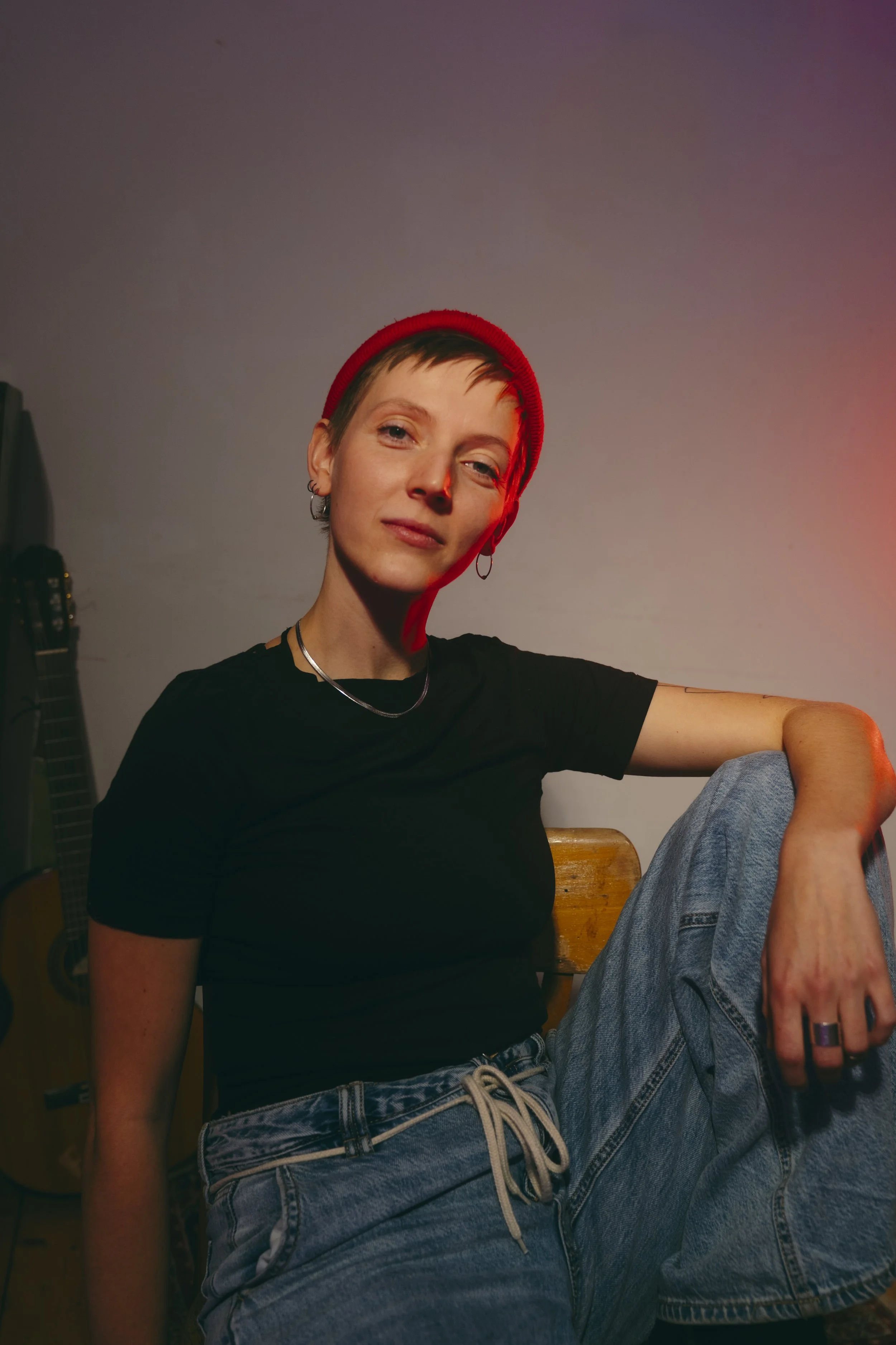 Lia L. Shoshann sitzend mit roter Mütze, im Hintergrund Gitarre, stolzer Blick, Arm lässig auf Bein abgestützt