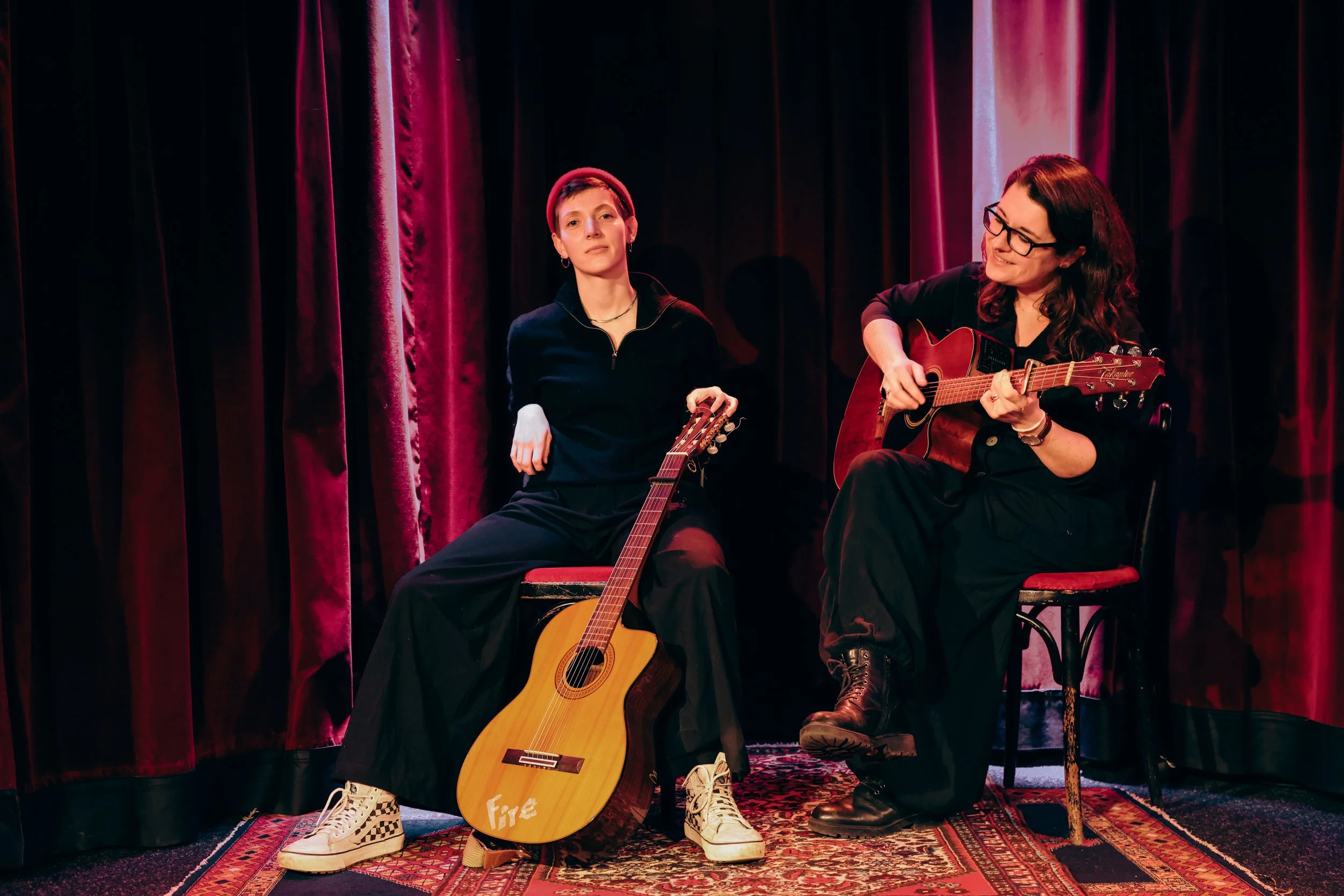 HANNA&LIA sitzend auf Stühlen vor schwerem roten Vorhang, Perserteppich. HANNA spielt Gitarre und sieht LIA and, LIA hält Gitarre, sitzt lässig und schaut nach vorn.