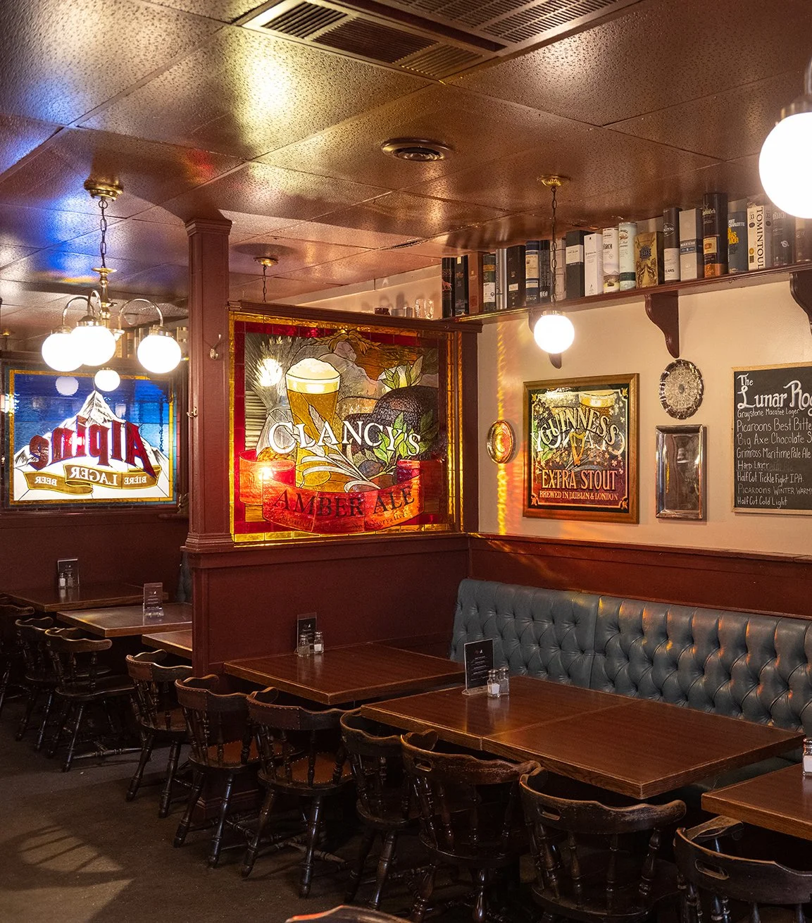 Interior of an Irish pub with wooden tables and chairs, upholstered seating, stained glass advertisements for Guinness and other beers, and a chalkboard menu on the wall.