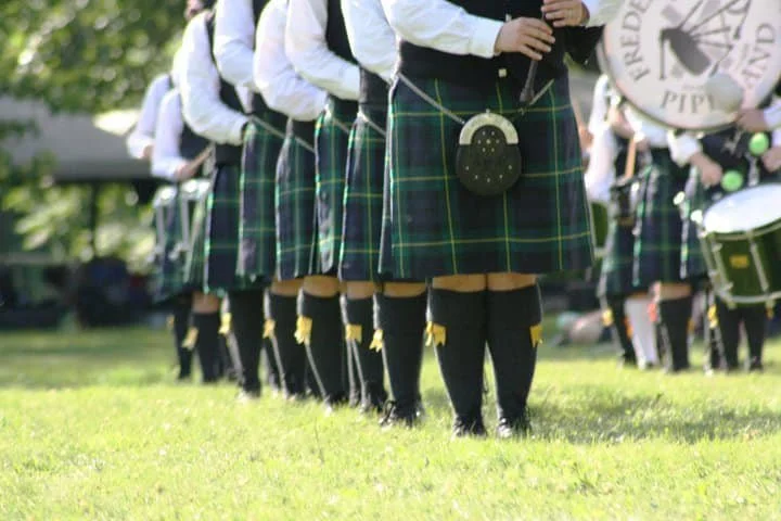 A line of people dressed in traditional Scottish attire, including kilts and black knee-high socks, standing on a grass field during daytime.