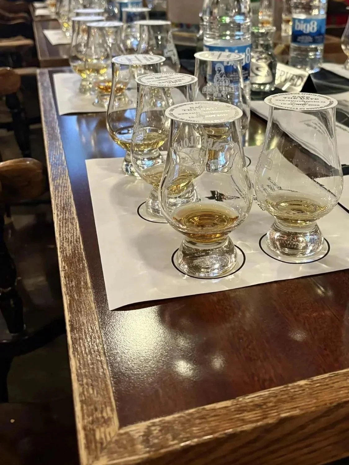 A table with several Glencairn glasses filled with small amounts of whiskey, arranged on a white tasting sheet inside a wooden tray in a bar or tasting room.