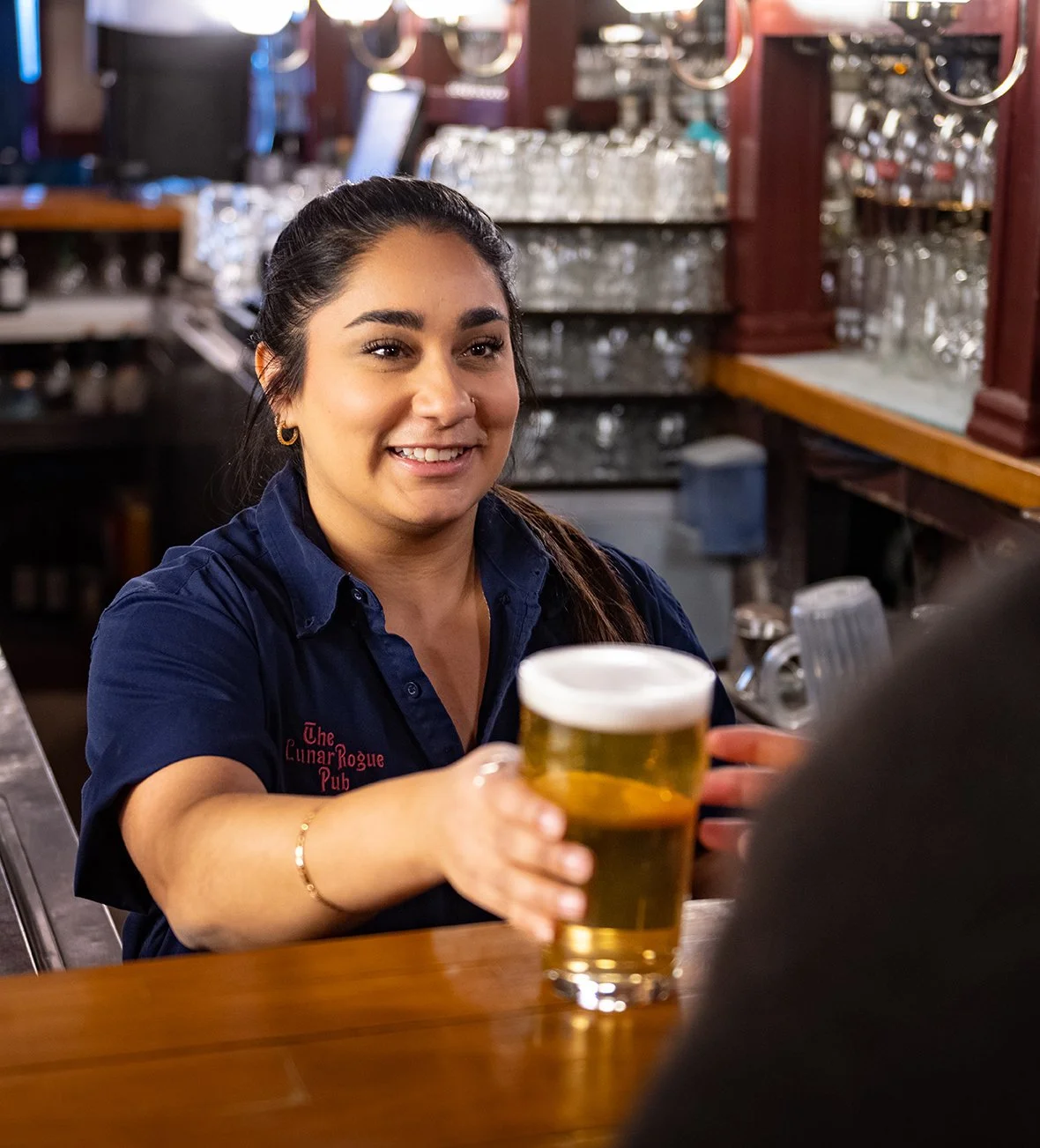A woman working as a bartender or server, smiling and handing a glass of beer to a customer in a pub or bar setting.