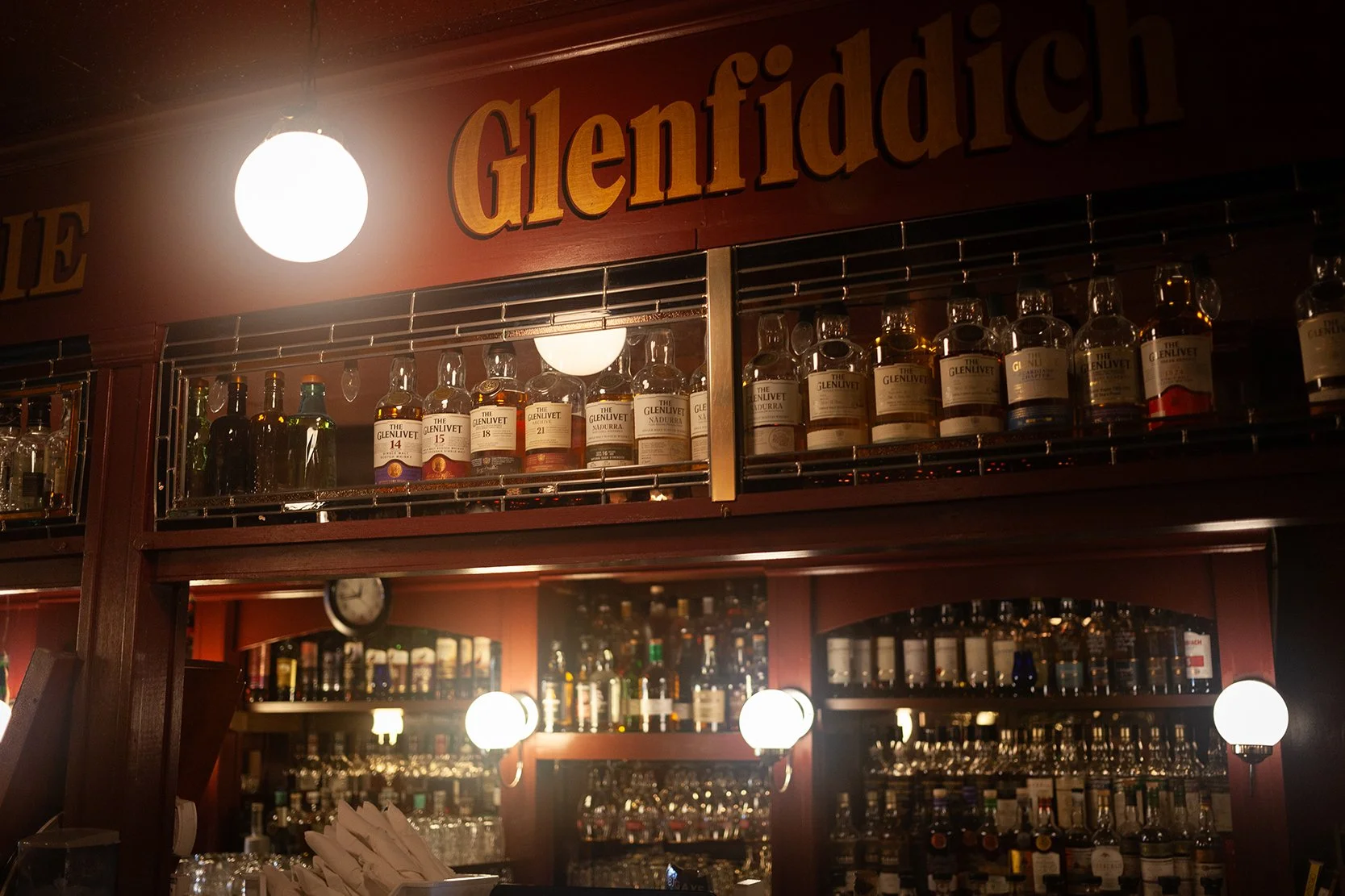 Bar with shelves of whiskey bottles, illuminated by round lights, and a sign reading 'Glenfiddich'.