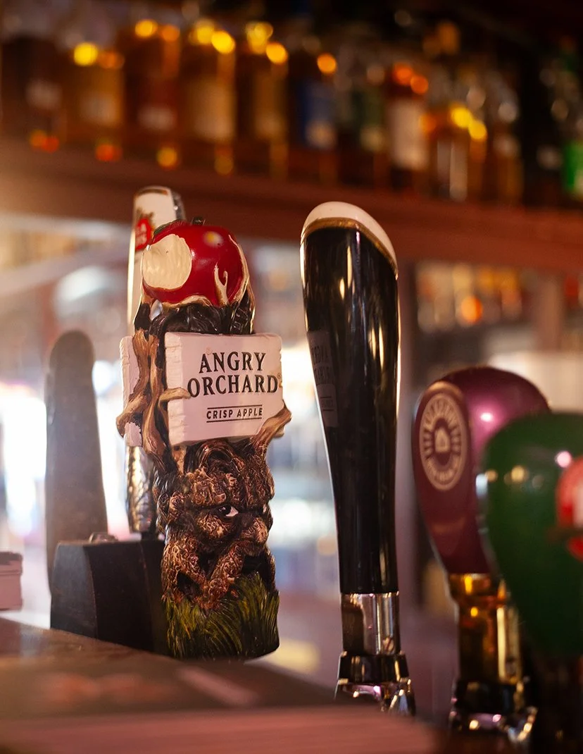 Close-up of beer taps, including one labeled 'Angry Orchard Crisp Apple,' in a dimly lit bar with blurred background of liquor bottles on shelves.