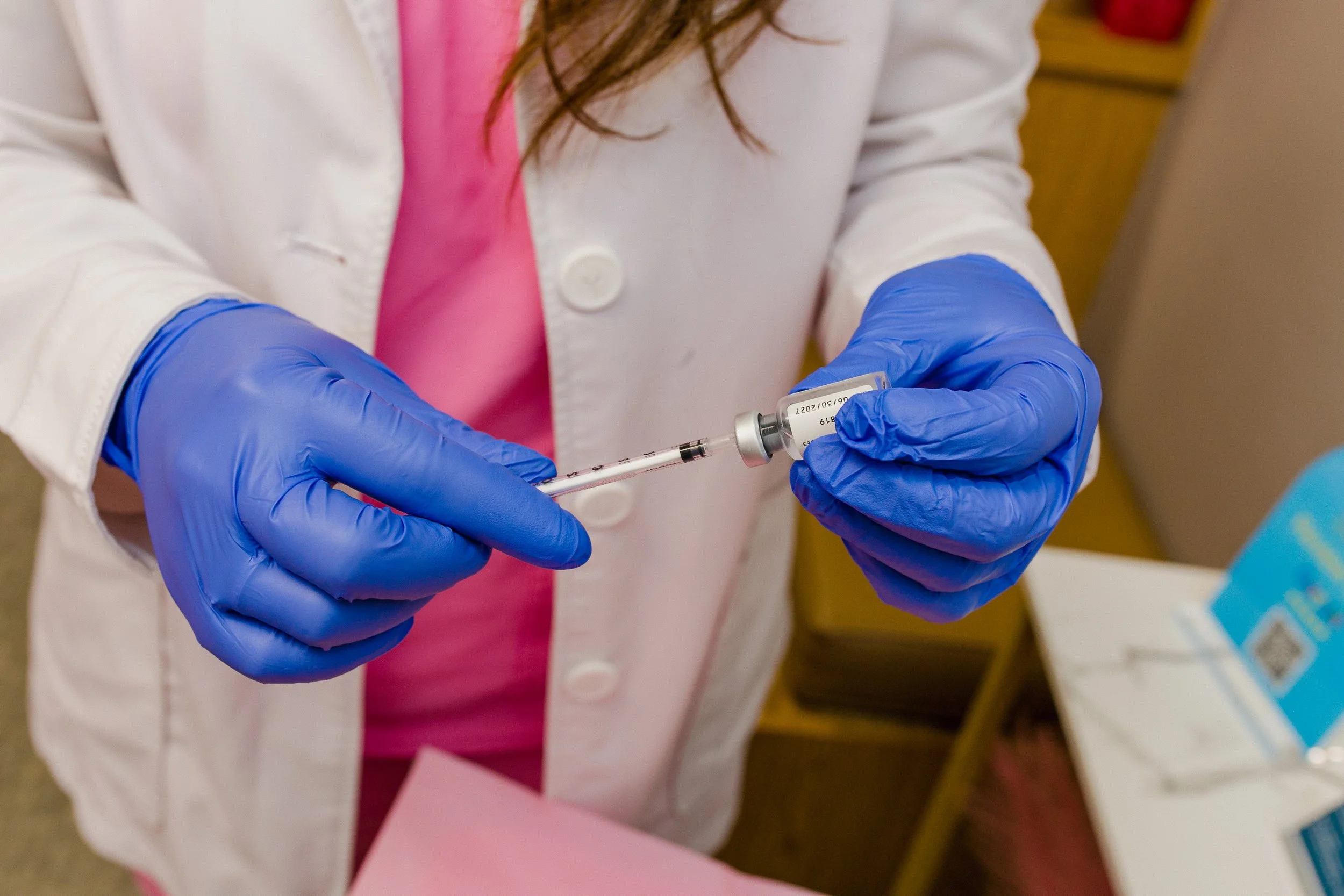 A healthcare professional in a white coat and blue gloves filling a syringe with a vaccine or medication from a small vial.