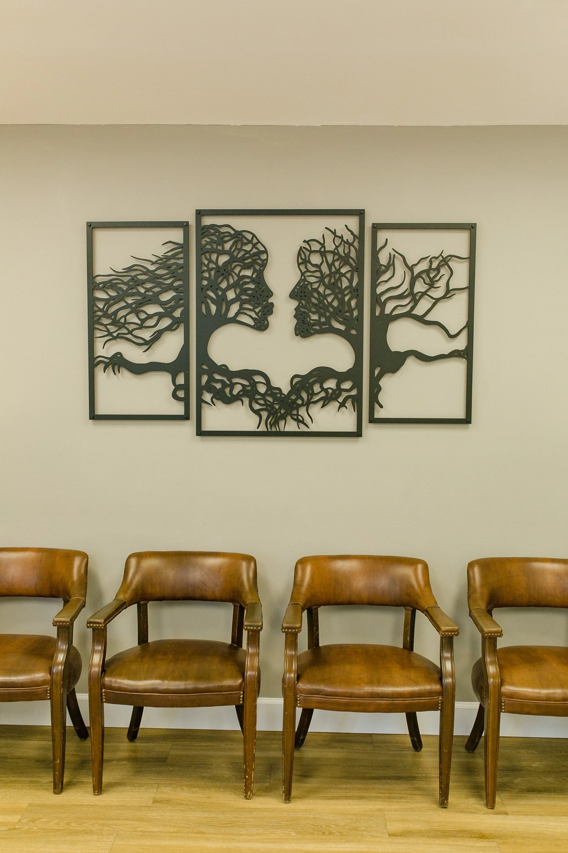 Decorative wall art depicting two faces in profile surrounded by tree branches, hung above four brown leather chairs in a waiting area.