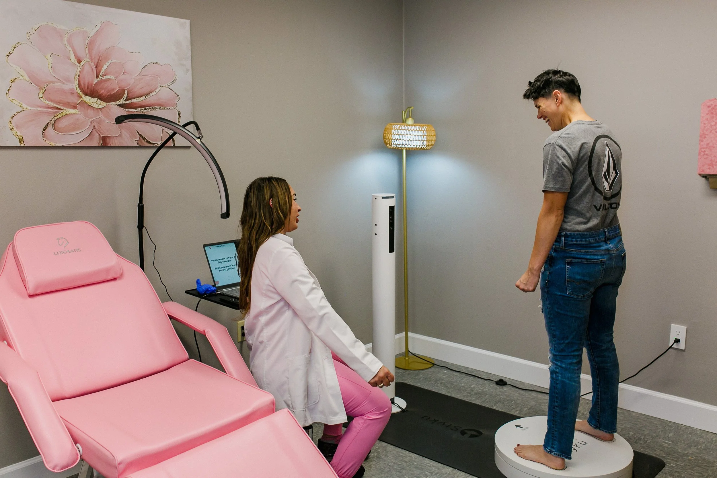 A woman is testing her balance on a digital force platform while a healthcare professional observes in a medical or wellness facility, with a pink treatment chair and a reading lamp in the background.