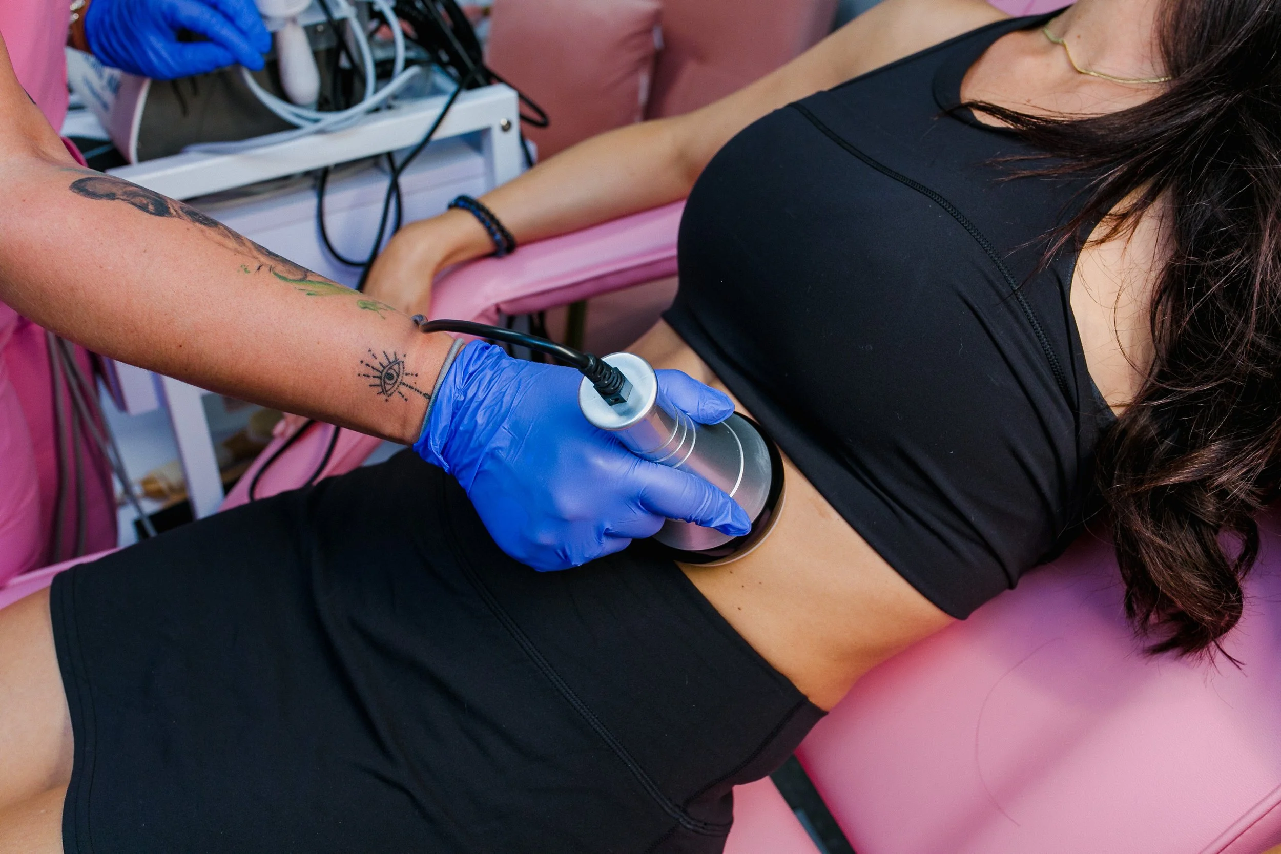 A woman receiving a cosmetic treatment with a handheld device on her abdomen while lying on a pink medical chair, with a technician wearing blue gloves.