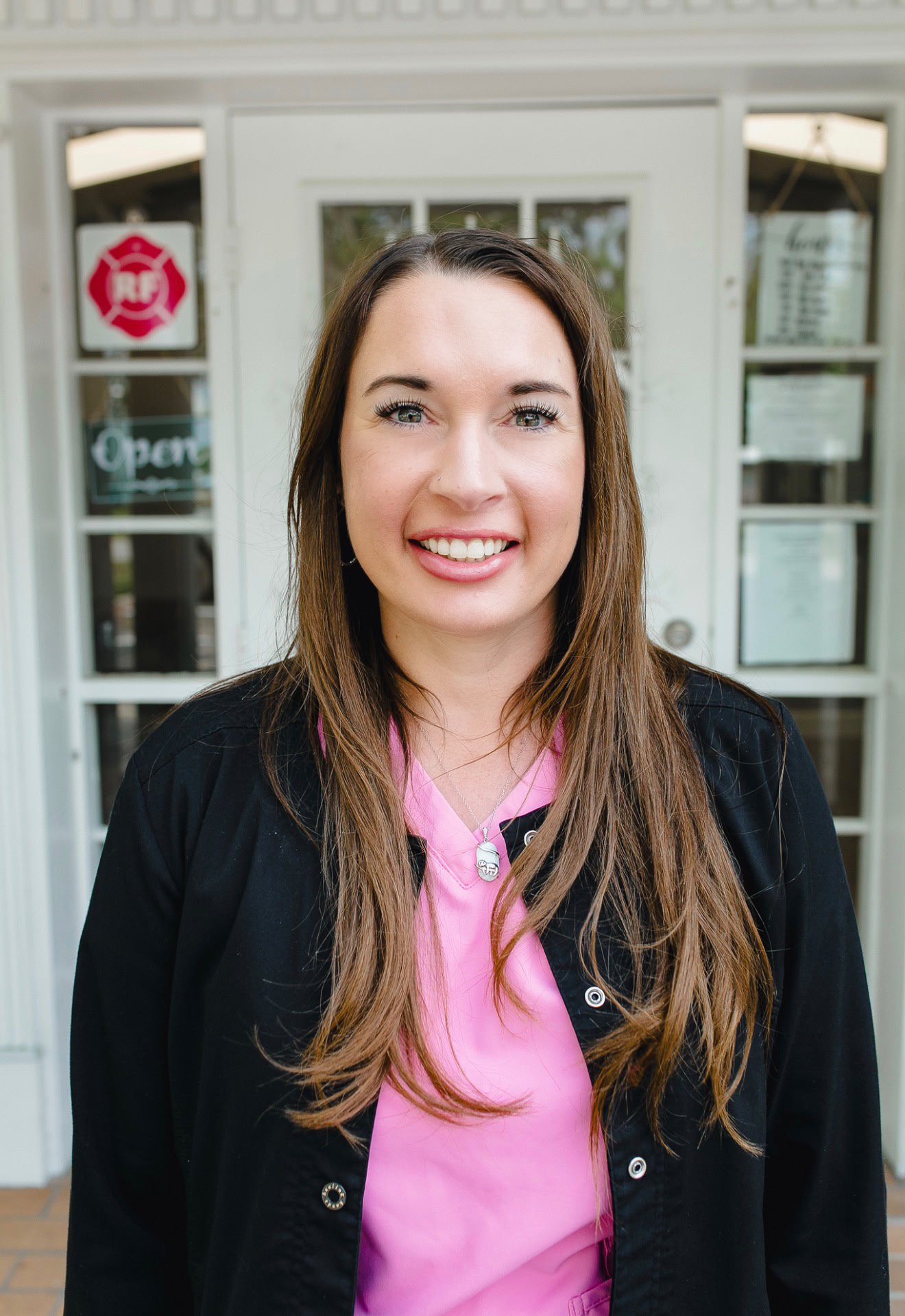 A woman with long brown hair smiling outdoors in front of a building with glass doors, wearing a pink shirt and black jacket.