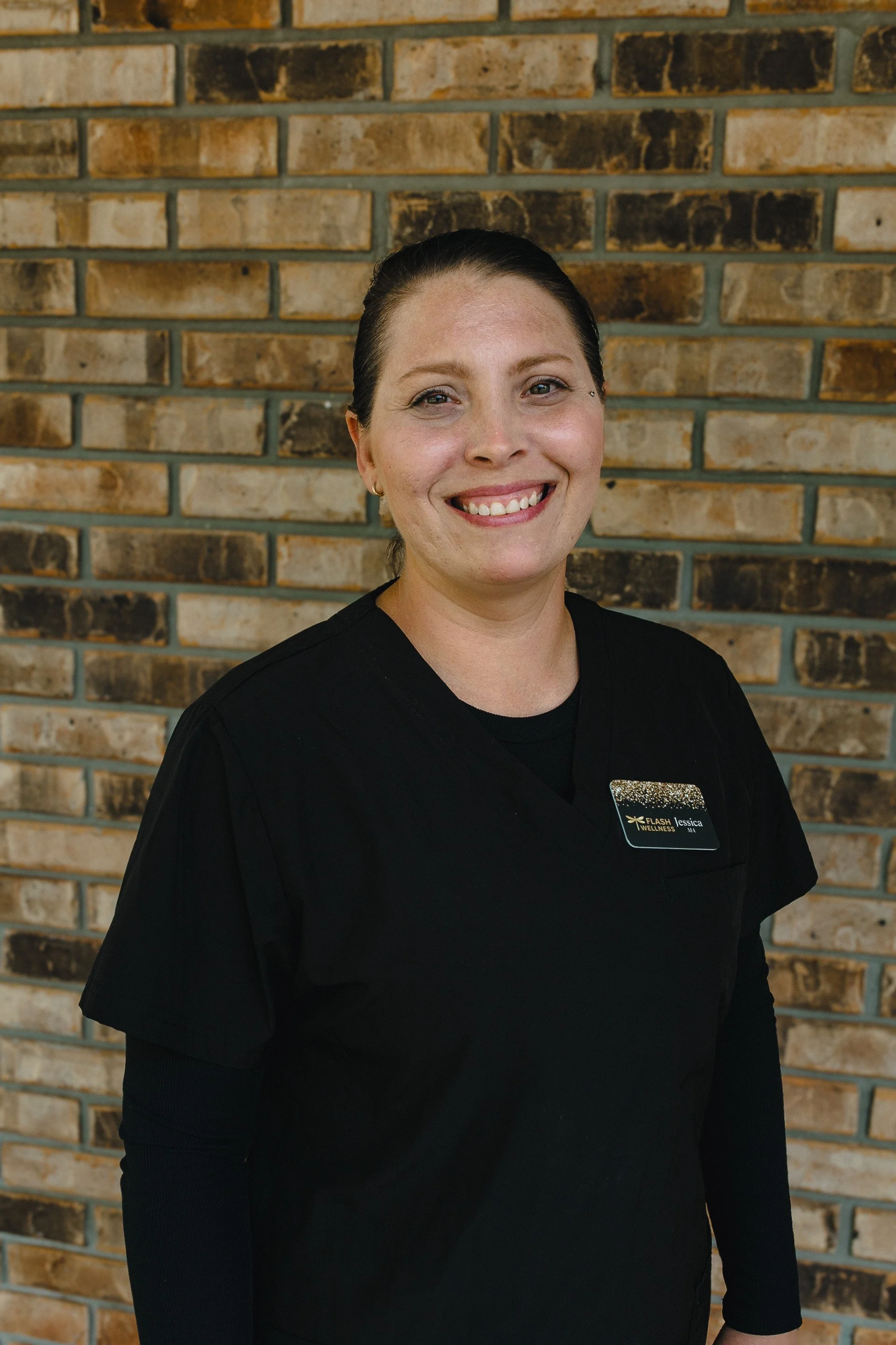 A smiling woman in a black uniform with a name tag that reads Jessica, standing in front of a brick wall.