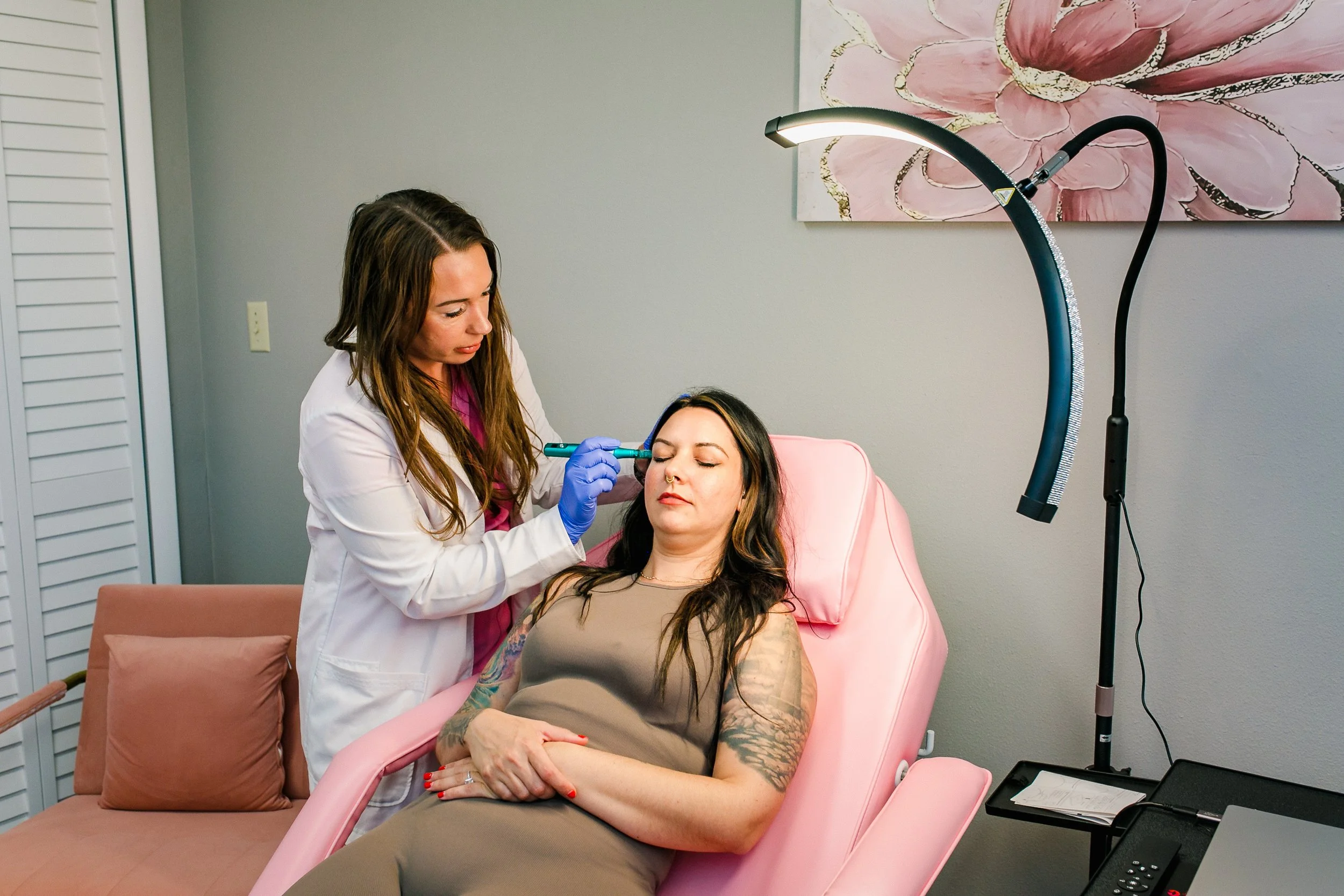 A woman with tattoos lying on a pink medical chair while a female doctor, wearing a white coat and blue gloves, administers a cosmetic treatment on her face using a pen-shaped device. The setting is a modern clinic with a gray wall, a pink abstract f
