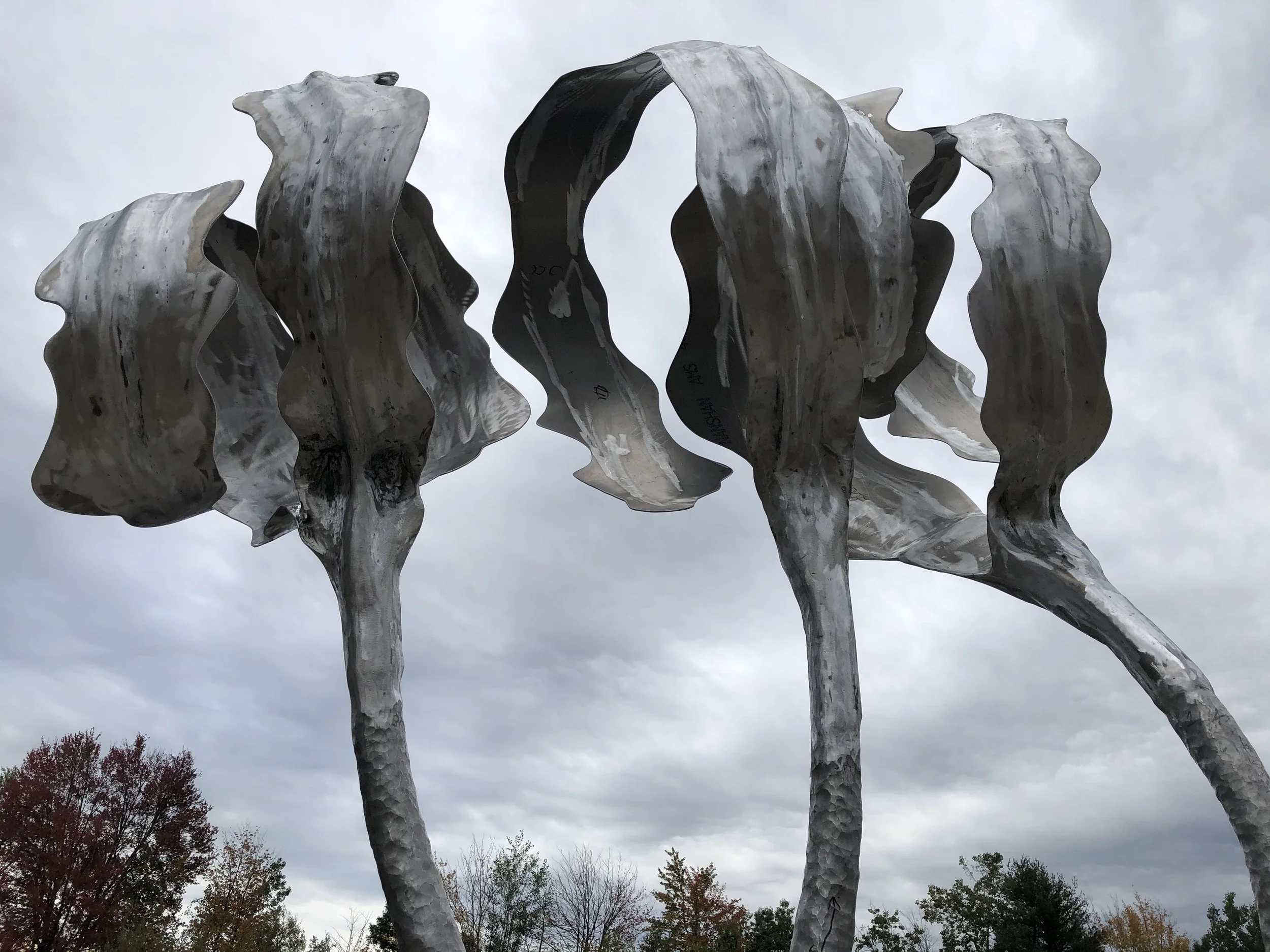 A large metal sculpture resembling a group of elongated, wavy, and curving stems of kelp, set against a cloudy sky and some trees in the background.