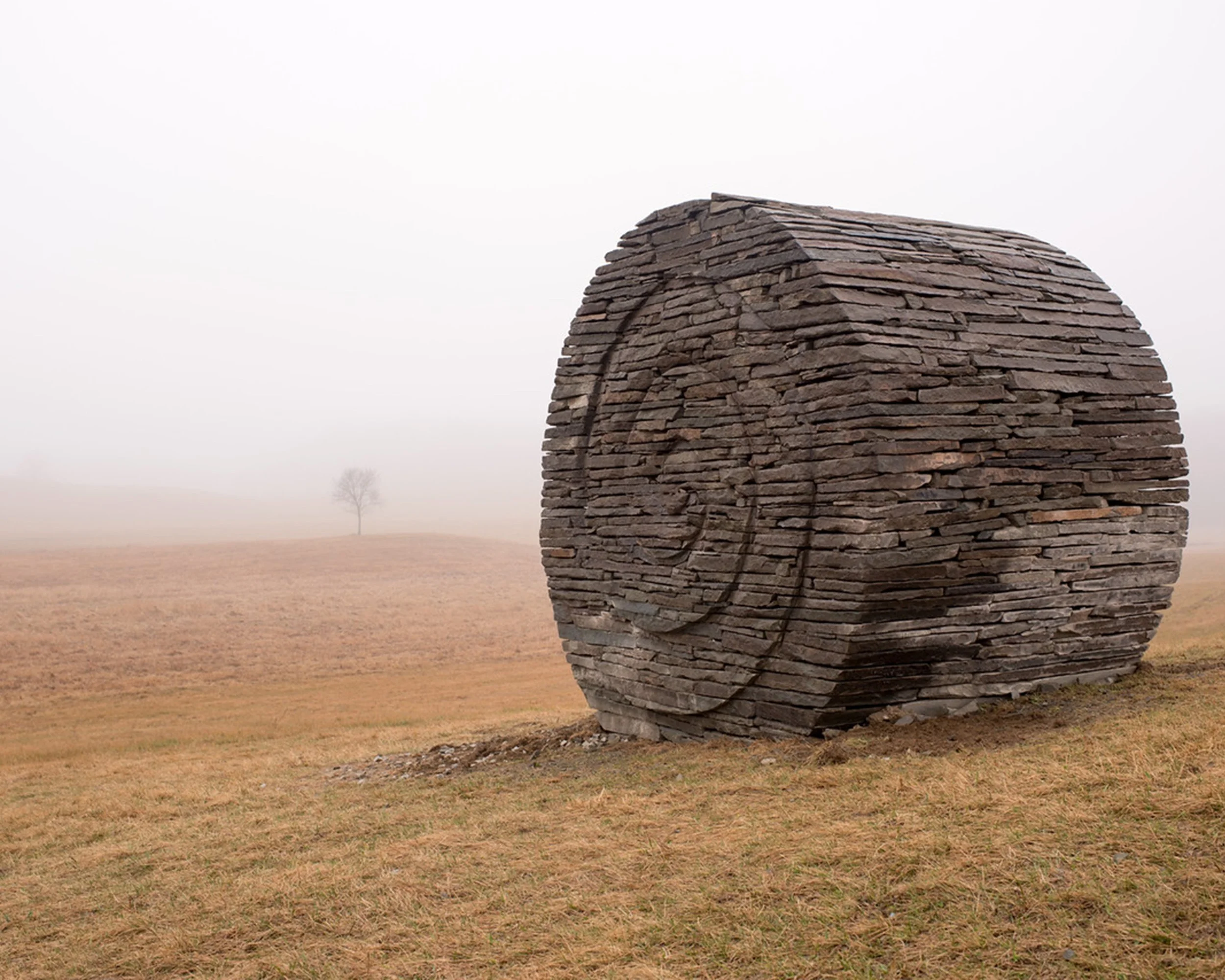 A large sculpture made of stacked stones depicting a round haybale, set in a foggy open field with a single distant tree.